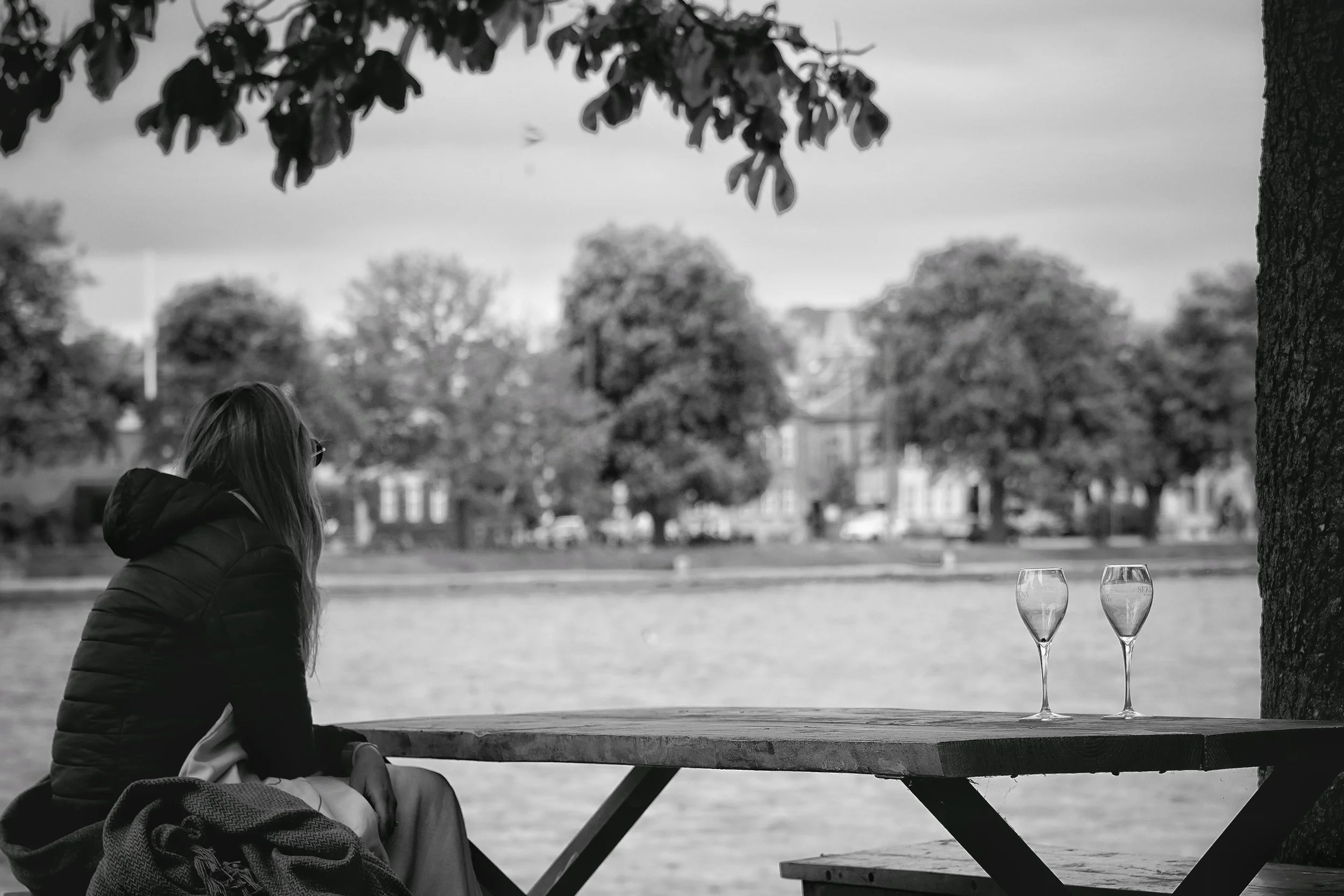 En kvinde sidder ved en træbord ved en sø med to tomme champagneglas, omringet af træer og bygninger i baggrunden.