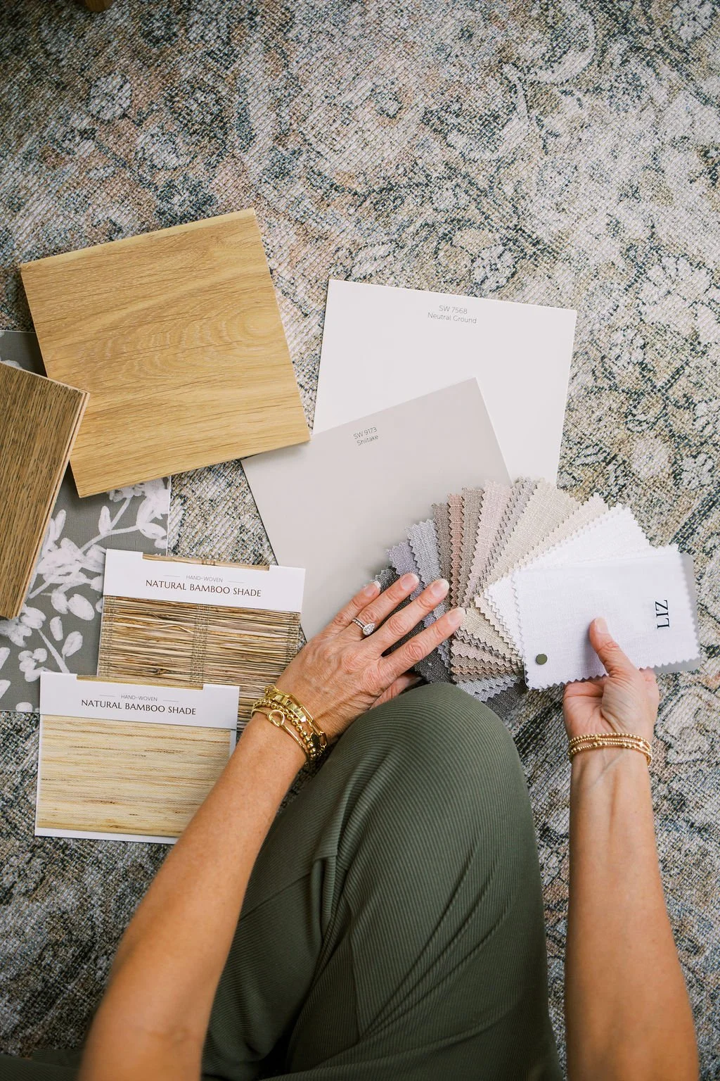 A person is sitting cross-legged on a patterned carpet, looking at several samples of flooring and fabric swatches. The samples include wood and bamboo textures, paint color swatches, and fabric samples, with the person's hands holding some of the swatches.