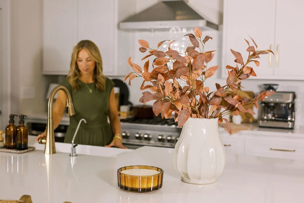 A woman in a green dress standing in a white kitchen with a large vase of brown and pink leaves on the counter.