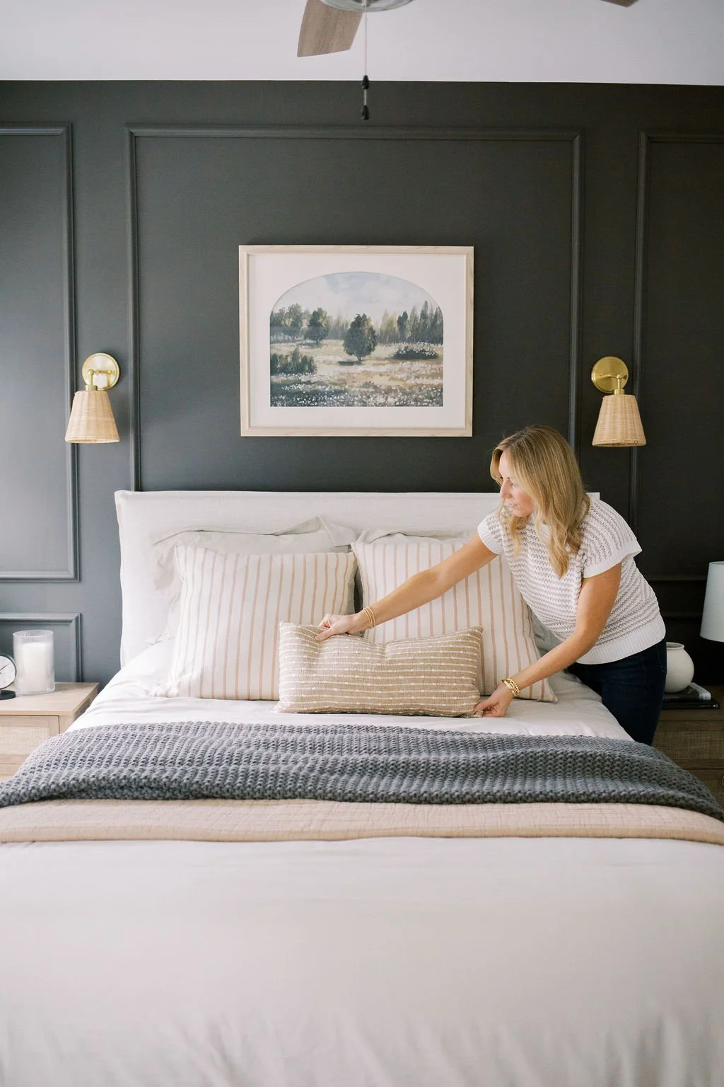 A woman making a bed in a bedroom with dark gray walls, white bedding, striped pillows, and artwork of a landscape above the bed.
