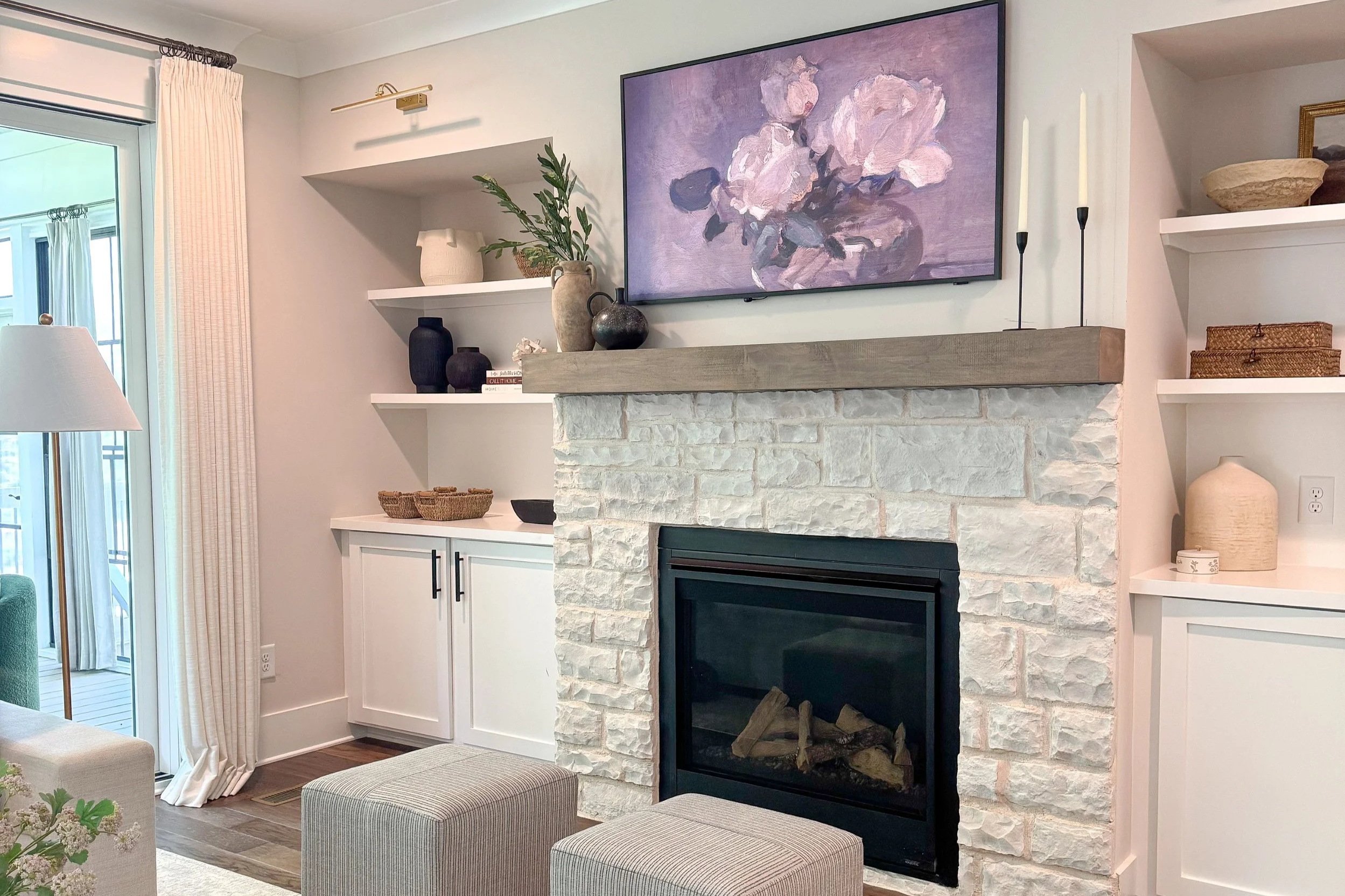 Living room with a fireplace, white stone surround, wooden mantel, wall-mounted TV with purple floral painting, white built-in shelves with decorative vases and baskets, sliding glass door with beige curtains, beige ottomans, and a chair with a white lampshade.