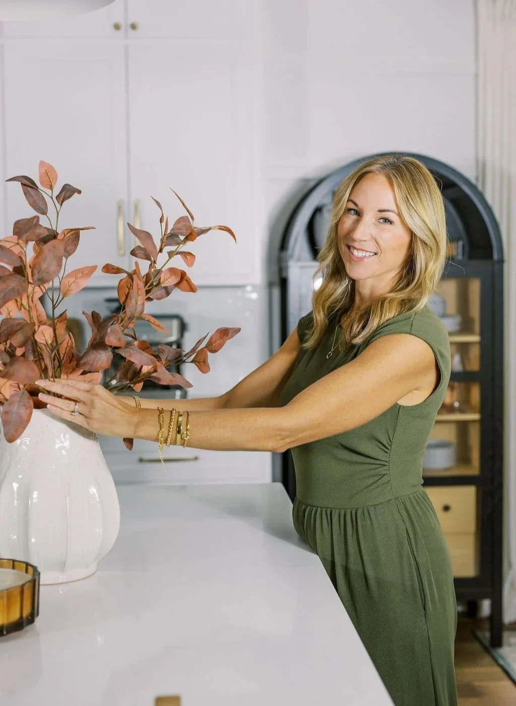 Woman arranging a large bouquet of reddish-brown leaves in a white vase in a modern kitchen.