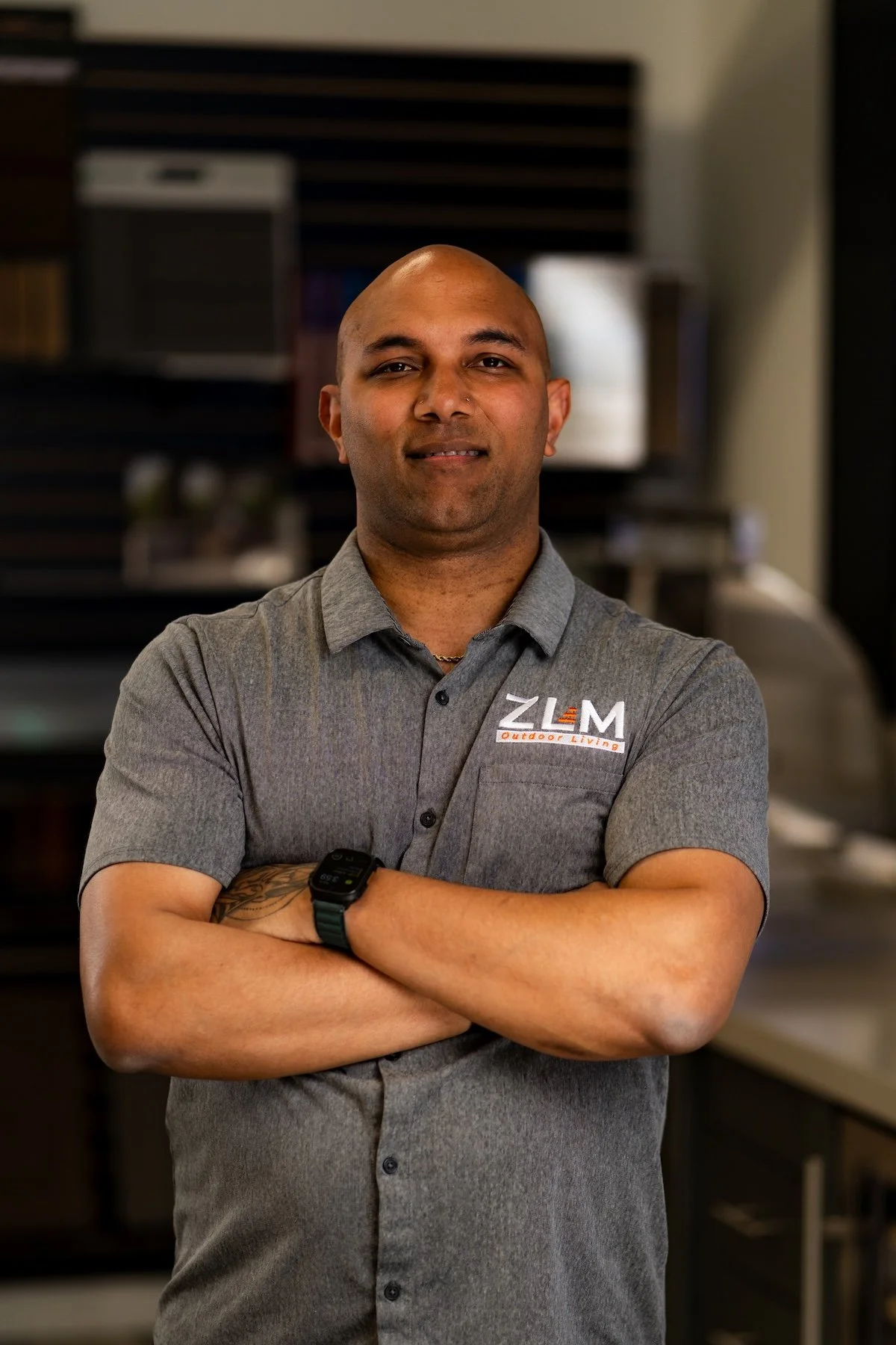 A smiling man with a shaved head is standing indoors with shelving and flooring samples in the background, wearing a gray short-sleeved collared shirt with the logo and text 'ZLM Outdoor Living'.
