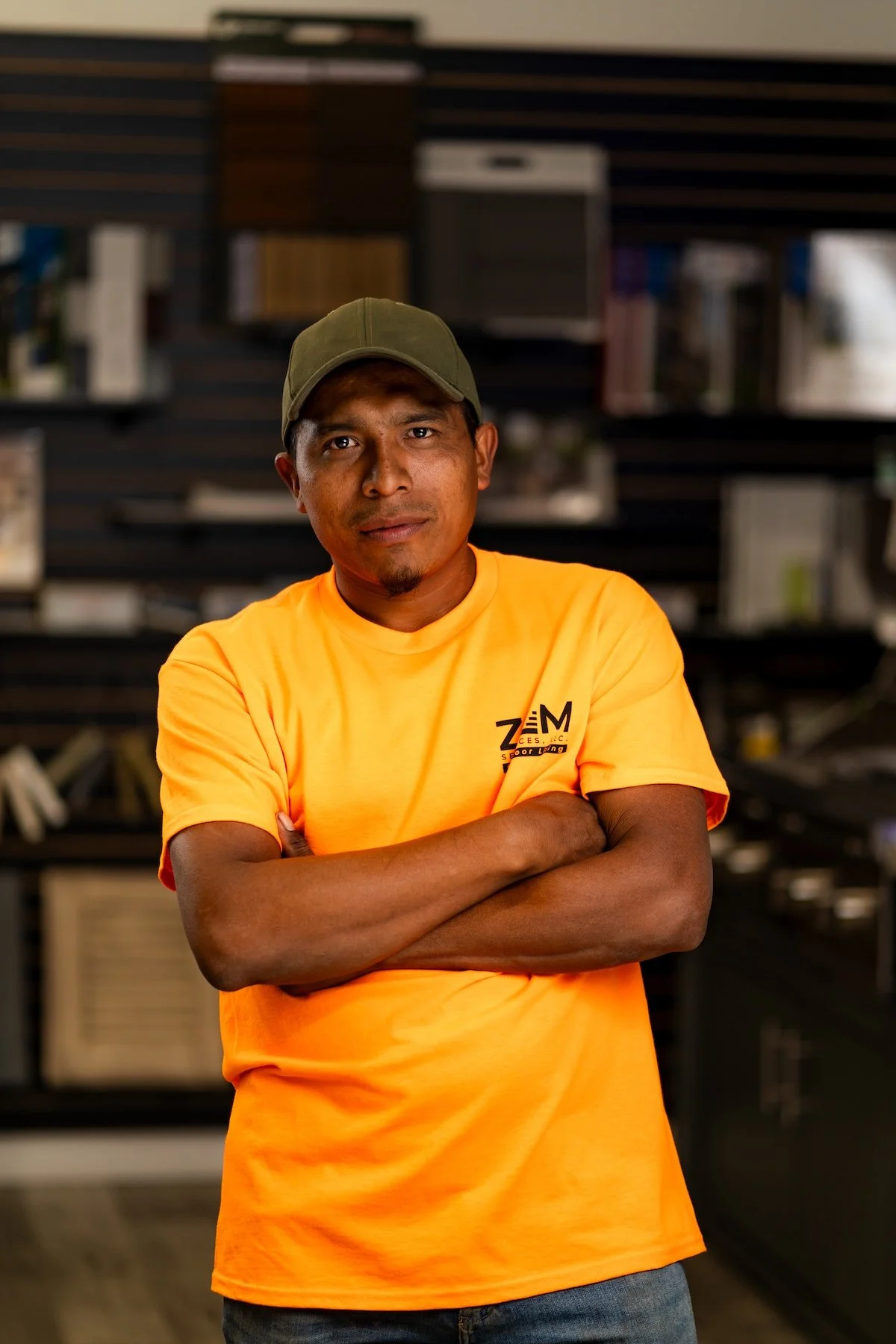 A man wearing an orange T-shirt and a camo baseball cap stands indoors with a neutral expression. The background is dark and features display racks of flooring samples.