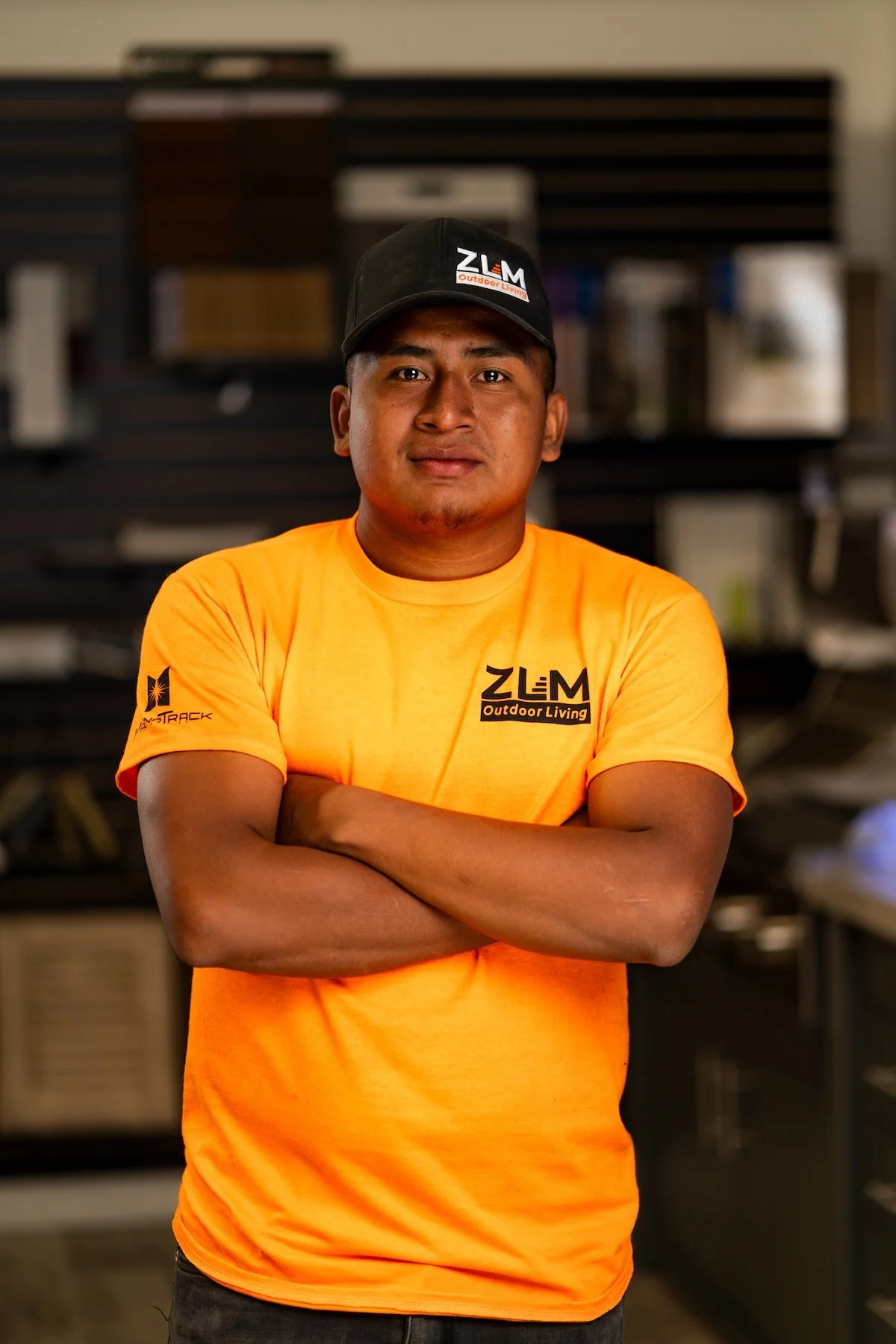 Young man wearing a blue LA Dodgers cap and orange ZLM Services LLC T-shirt standing inside a hardware or flooring store.