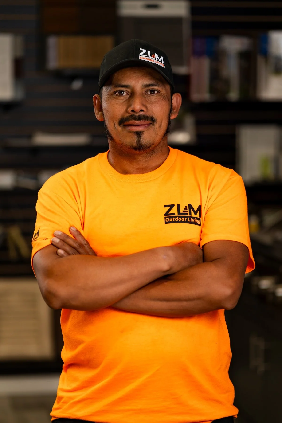 Man with black hair, mustache, and goatee wearing an orange T-shirt and black cap in a workshop or hardware store setting.
