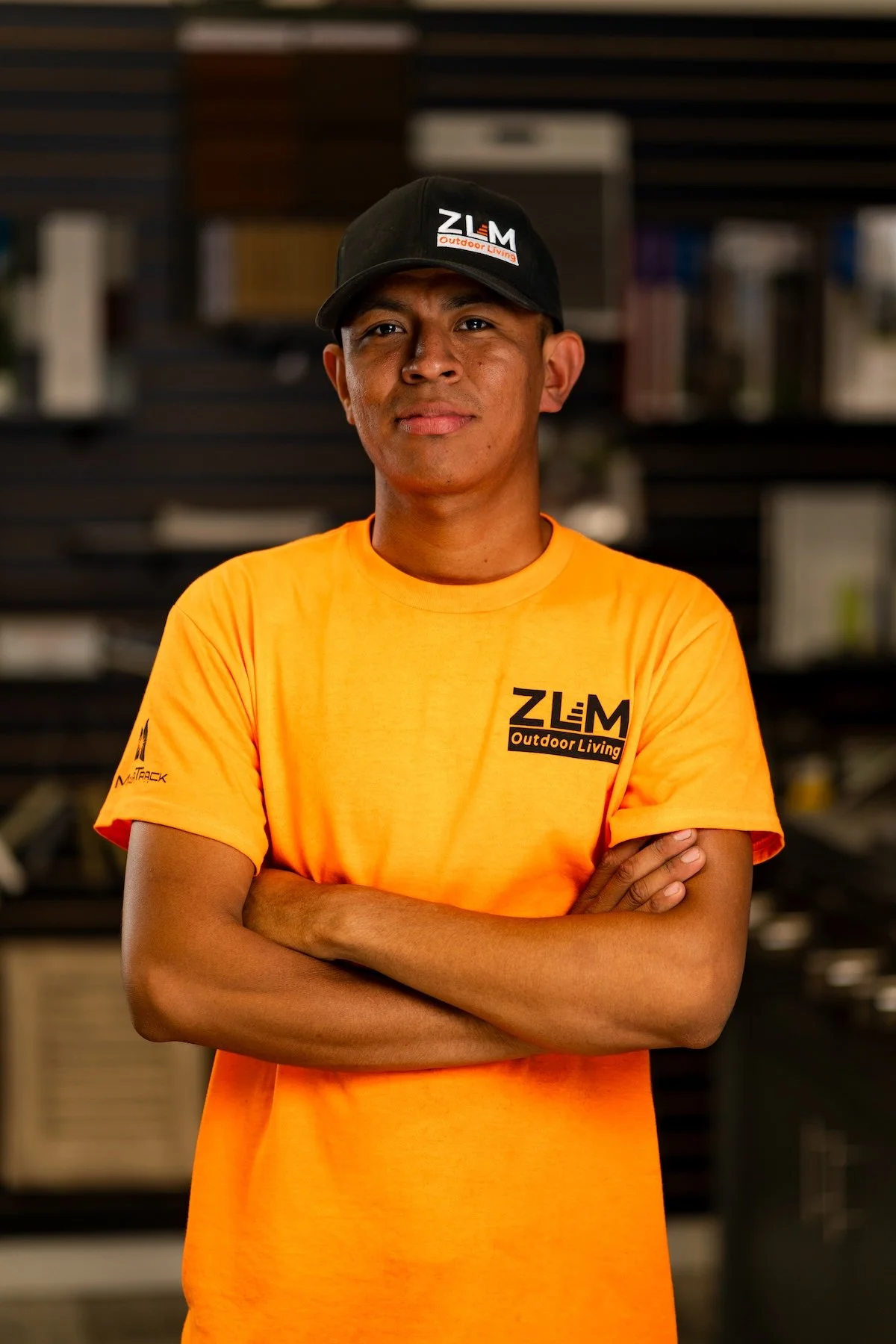 A young man wearing a red cap and a black hoodie with an orange logo stands indoors with a dark background and shelves in the background.