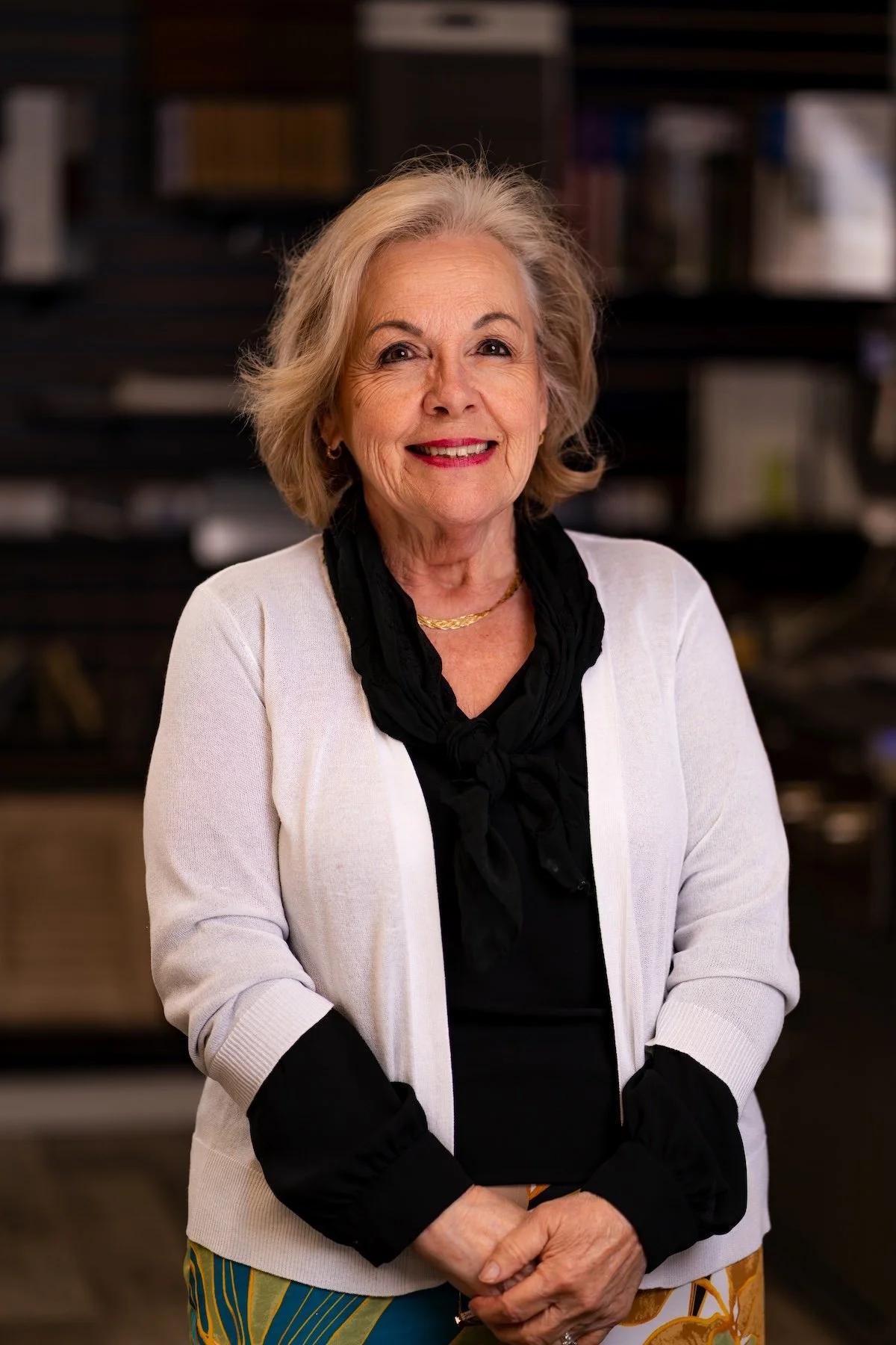 A smiling older woman with gray hair wearing a blue cardigan, floral blouse, pearl necklace, and a white wristwatch, standing in a store with wood flooring and carpet samples in the background.