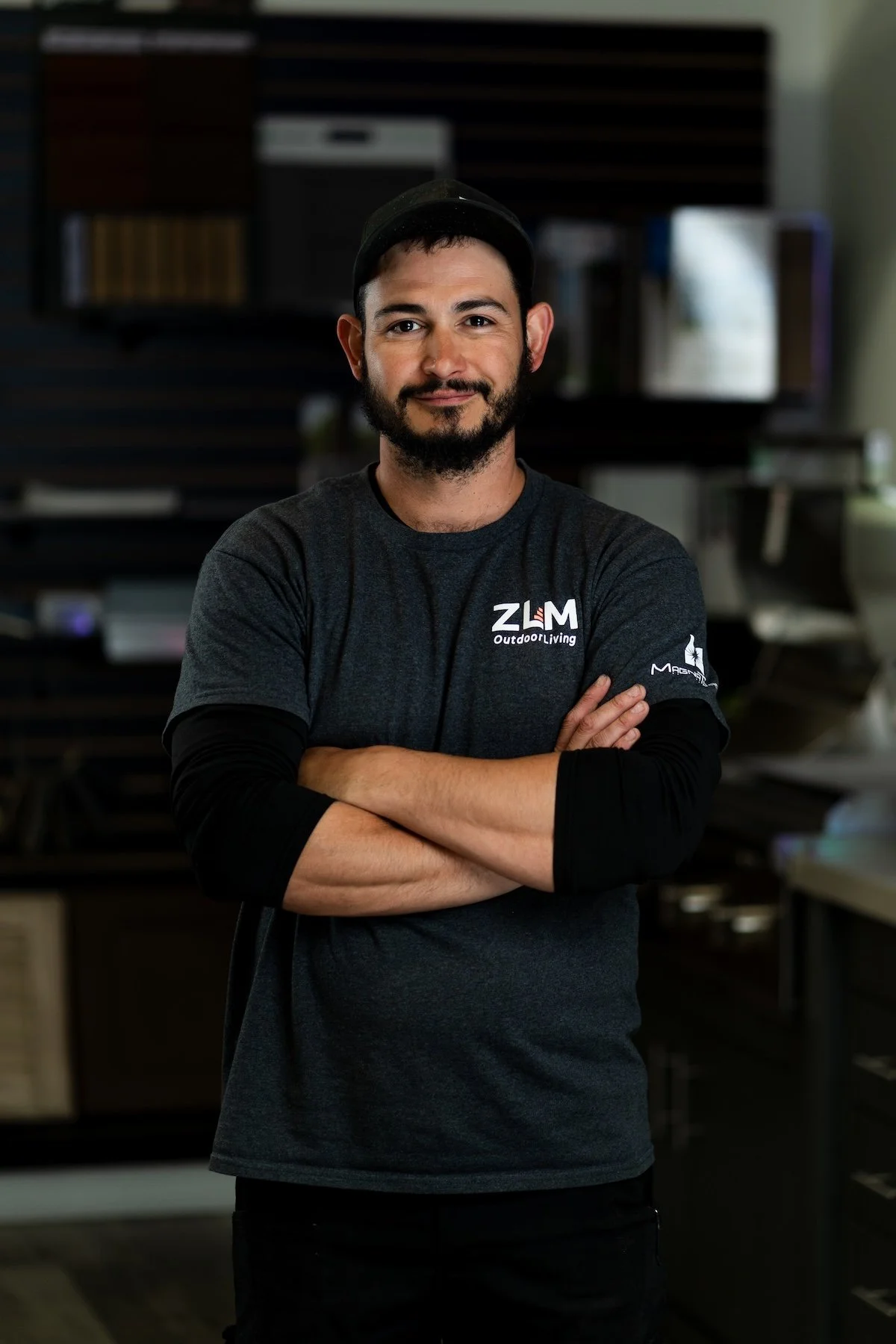 A man with a beard and tattoos wearing a gray polo shirt with ZLM Services LLC logo, standing in a store with flooring samples in the background.