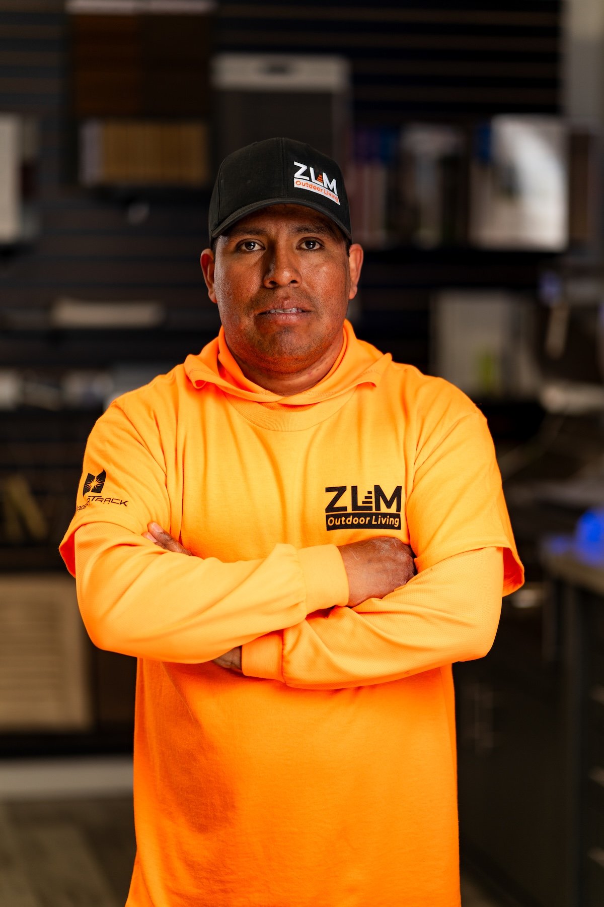 A man with dark hair, wearing a blue baseball cap backwards, an orange long sleeve shirt with black text and logo, standing indoors with a blurred background of shelves.