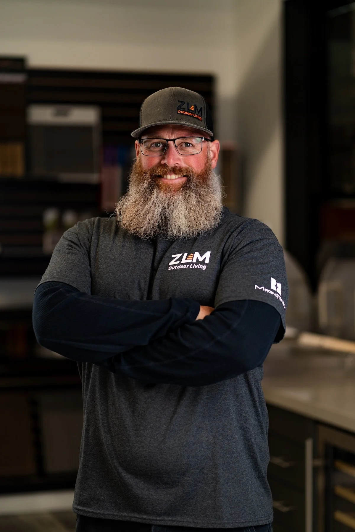 A middle-aged man with a thick beard and glasses, wearing a dark gray polo shirt and a black cap, stands in an indoor retail or hardware store. The store background includes shelves with various products and a large advertisement featuring two people seated on a white couch.