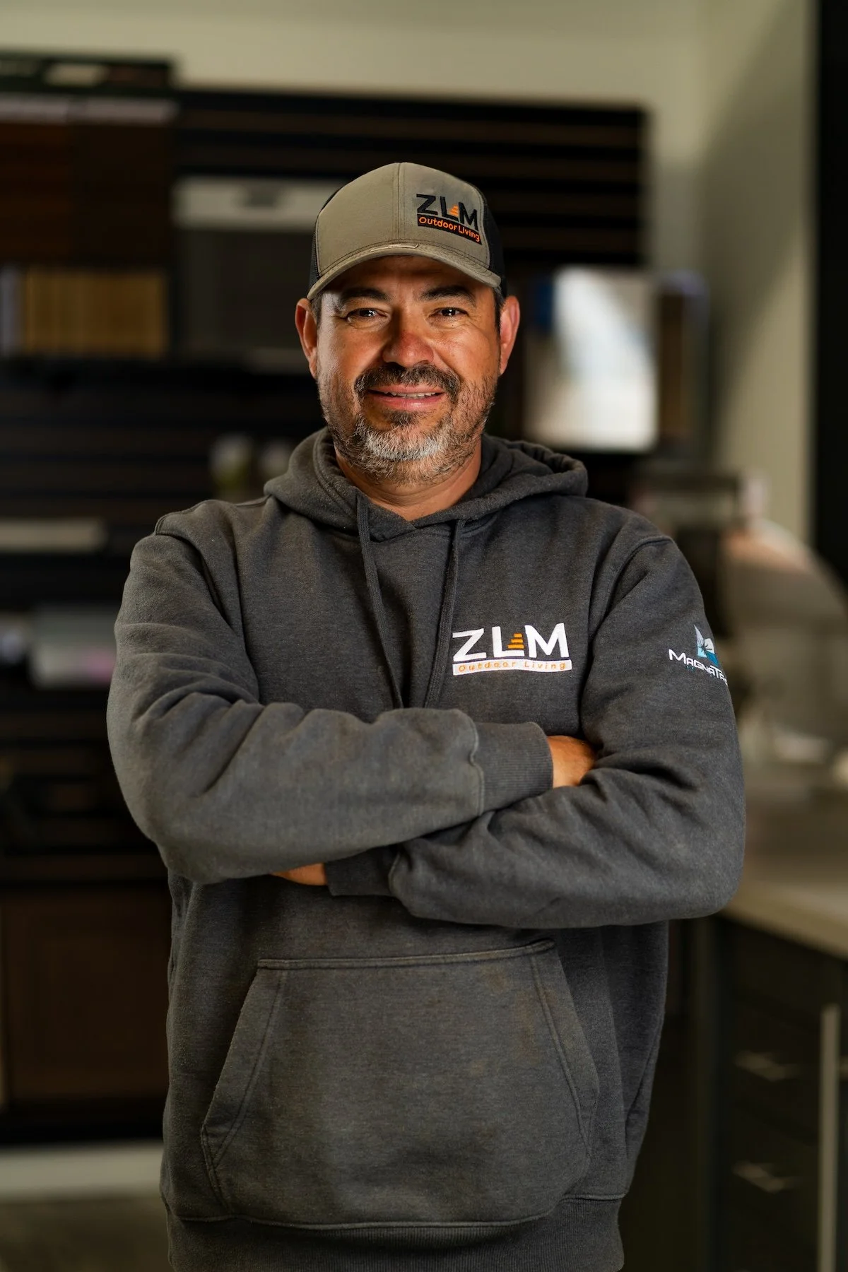 Man wearing a black hoodie and a baseball cap, standing indoors with a smile, shelves with samples in the background.