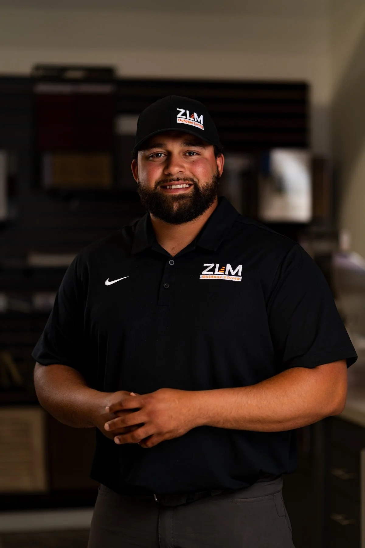 A smiling man with dark curly hair and a beard, wearing a gray polo shirt with an embroidered logo that says "ZLM Services LLC" and "Outdoor Living," standing with arms crossed inside a store surrounded by samples of wood or flooring.