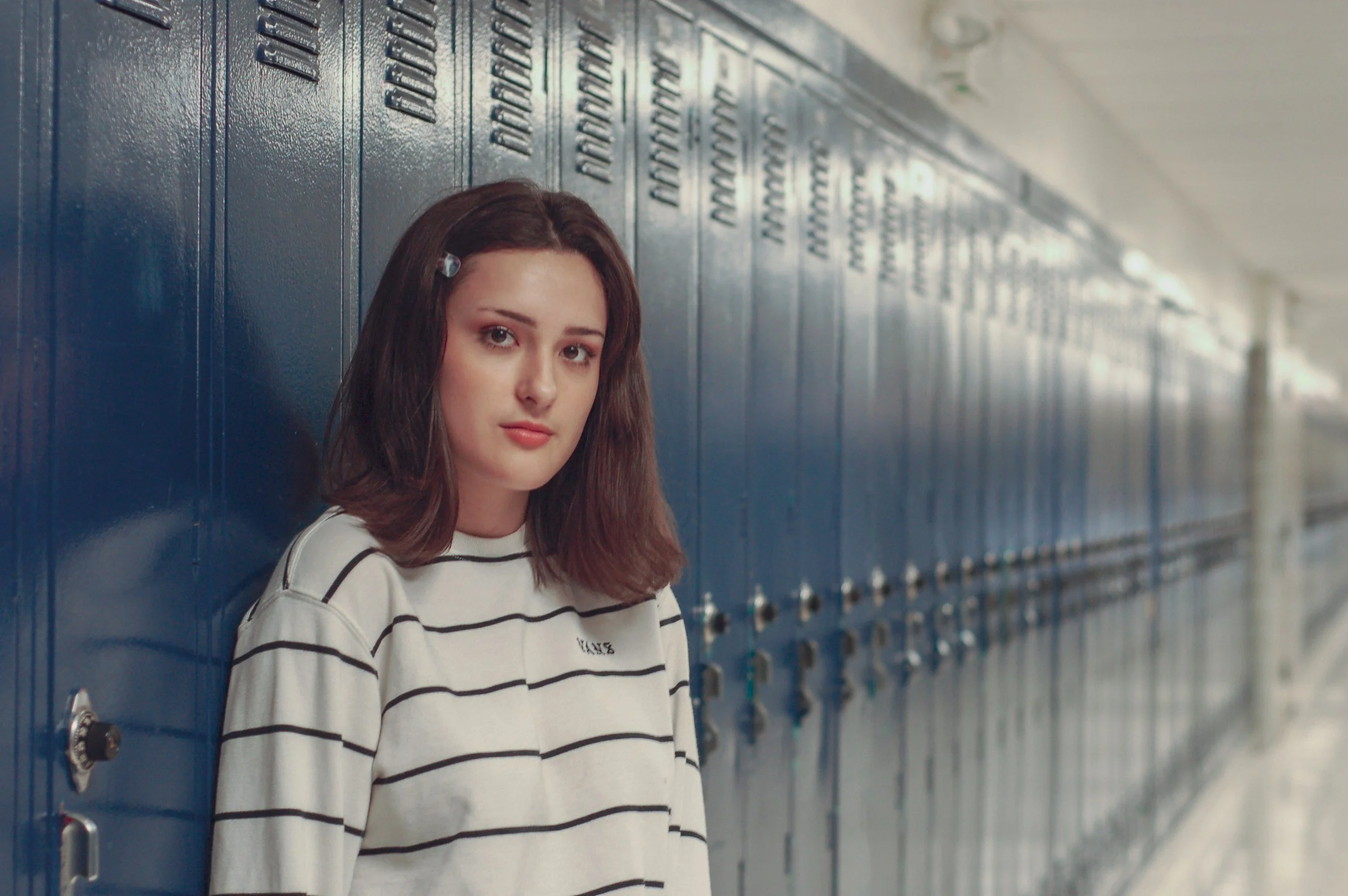 A teenage girl with shoulder-length brown hair, wearing a white and black striped shirt, leaning against blue lockers in a school hallway.