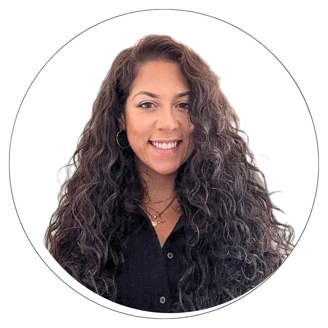 A woman with long brown hair, wearing glasses, earrings, and a gold necklace with a pendant, smiling at the camera against a white background.