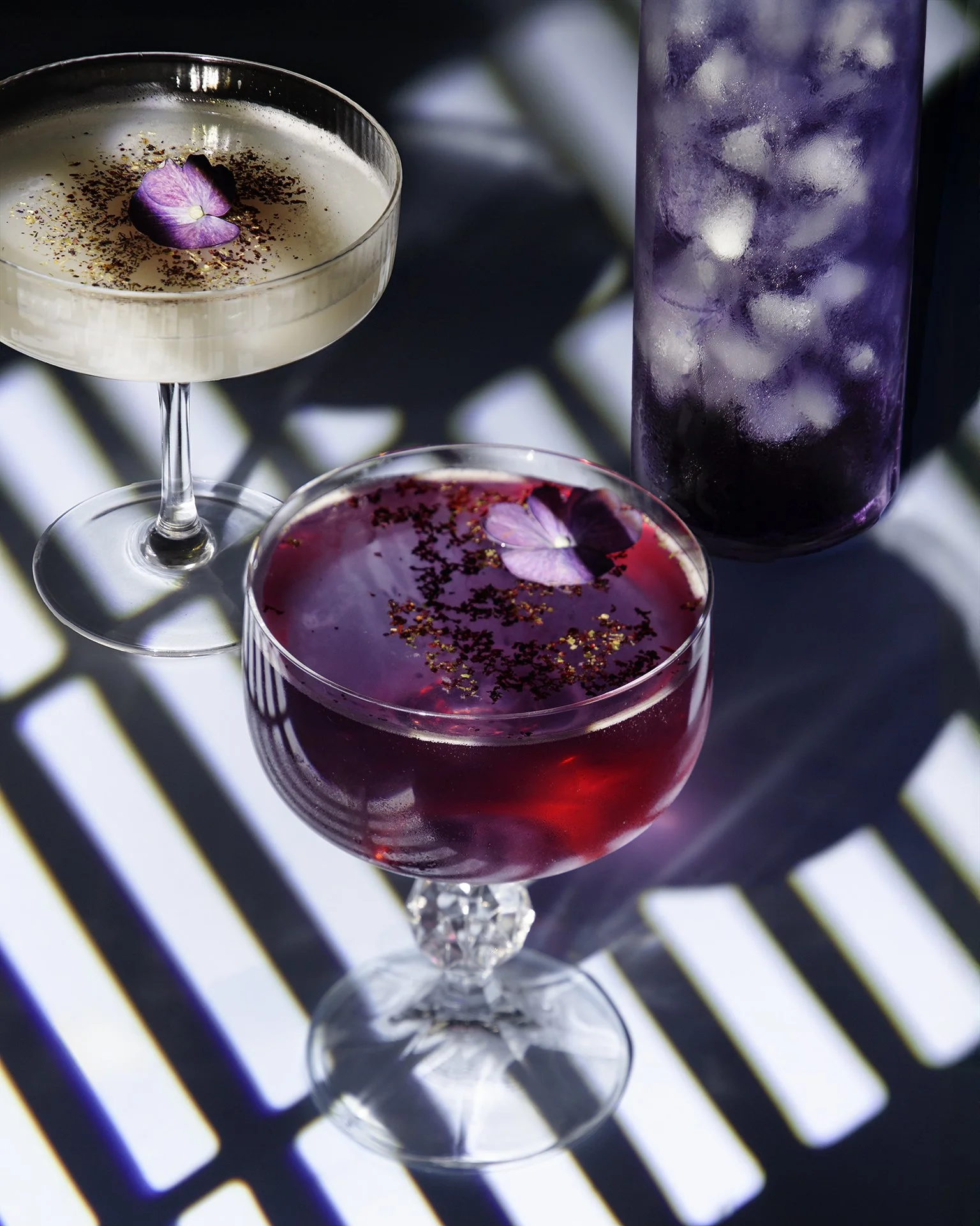 Two cocktail glasses with purple and pink drinks, garnished with edible flowers and sprinkled with red seasoning, on a black surface with striped shadows.
