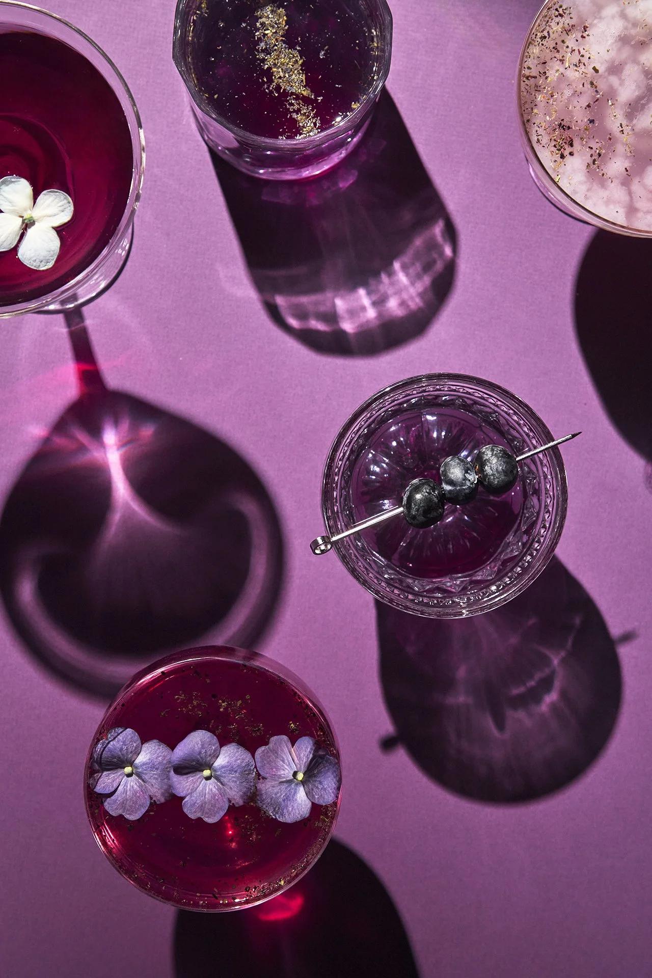 Several colorful cocktails on a pink surface with floral garnishes, garnished with flowers and berries, casting vibrant shadows.