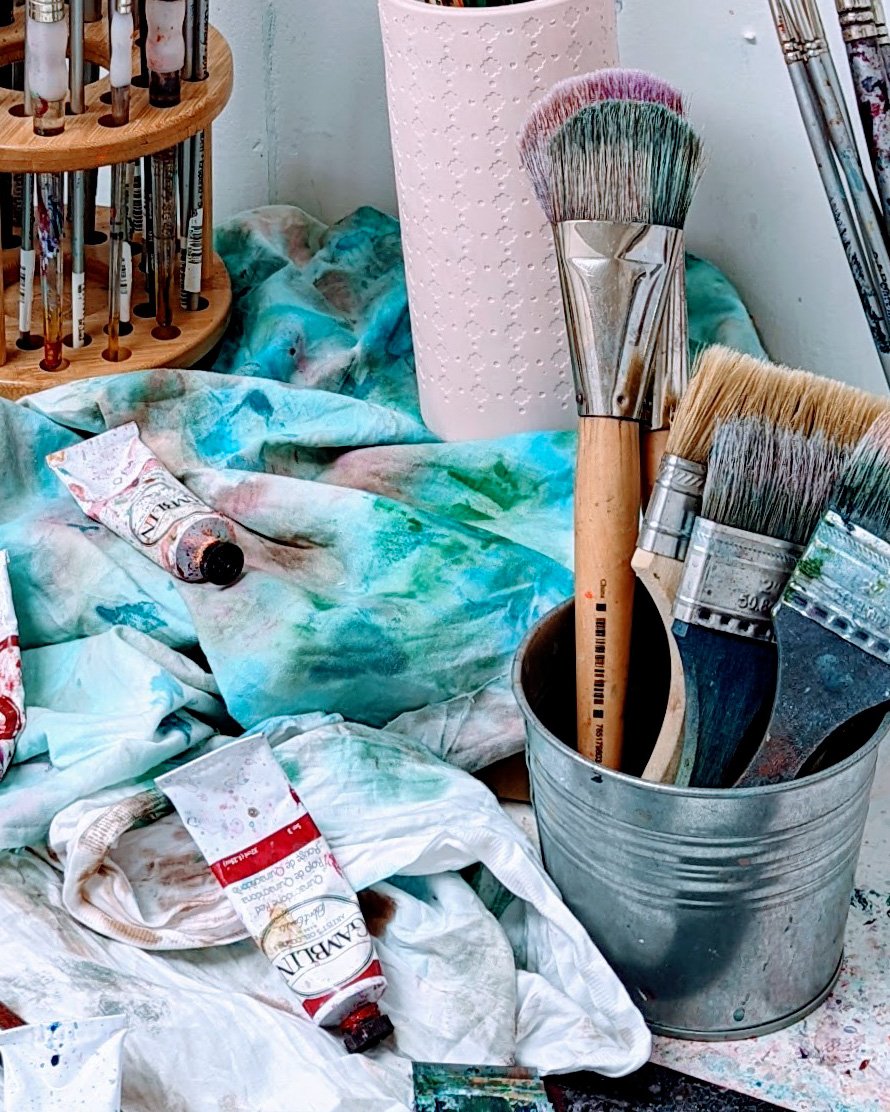 Paint brushes in a metal container, spilled artistic paints and tubes on a stained cloth, and a paint tube on a stand with other art supplies.