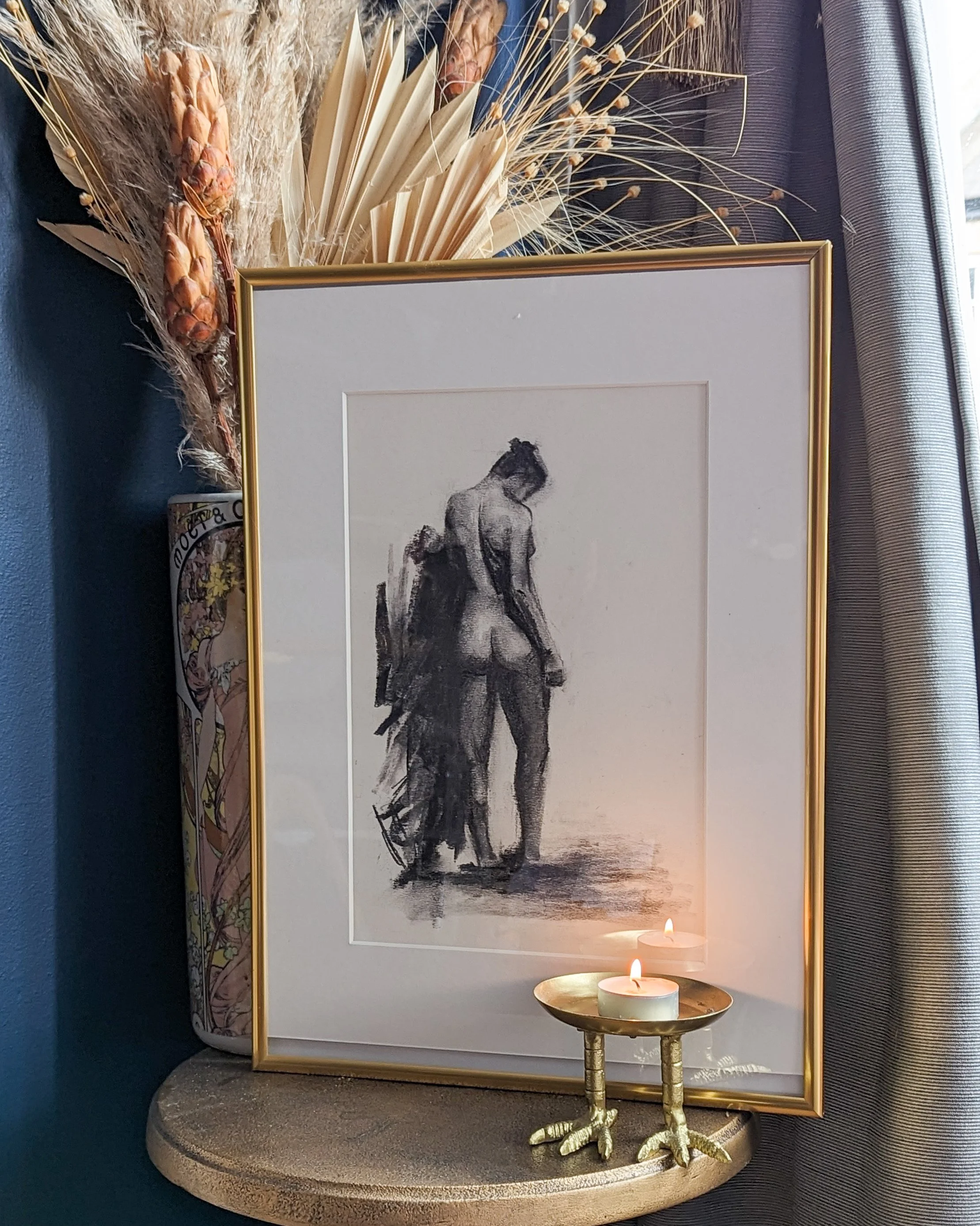 A framed black and white sketch of a nude woman from behind, with her hair up, standing on a flat surface. The frame is gold. In front of the frame, there is a small gold candle holder with three lit tealight candles shaped like bird feet, on a round table. In the background, there are dried flower and plant arrangements on the left, and a window with curtains on the right.