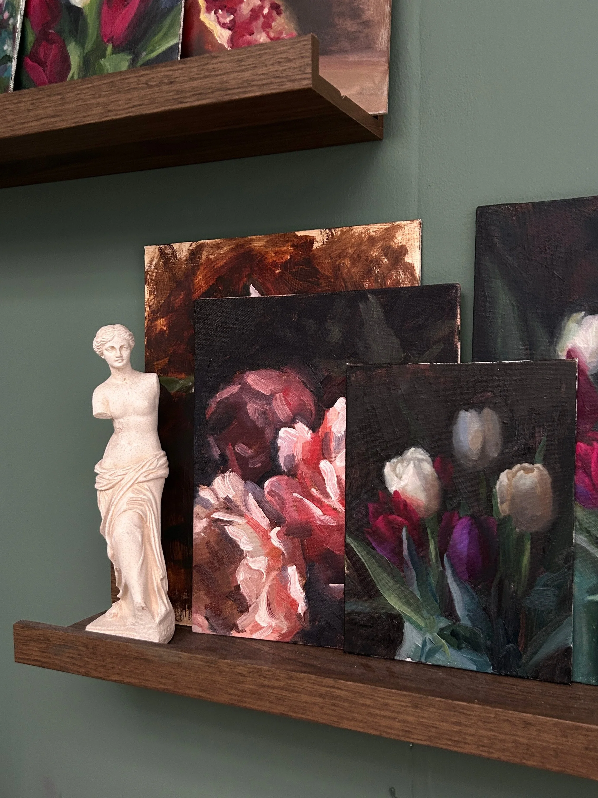 A wooden shelf holding several small paintings with floral themes and a white sculpture of a woman.