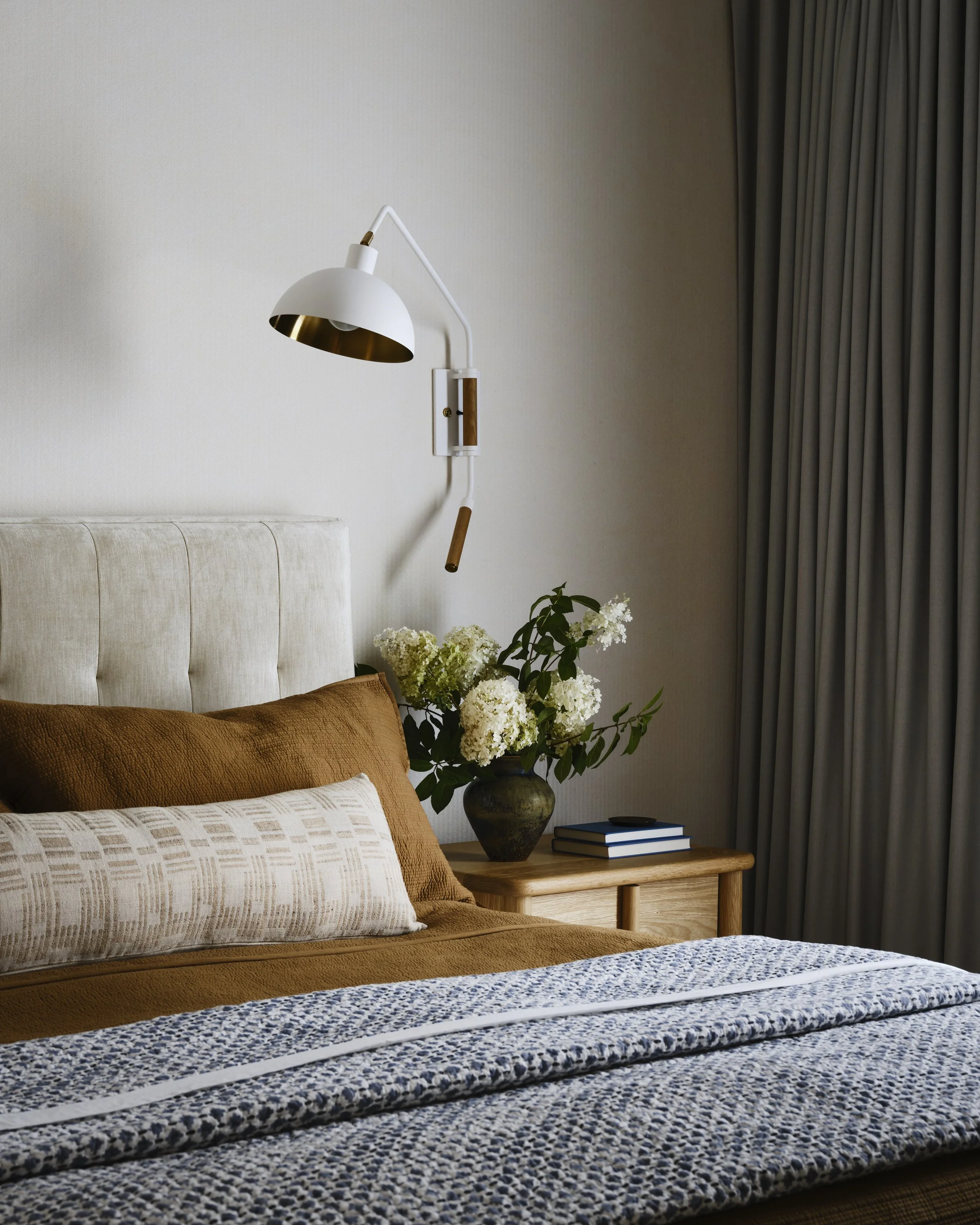 A bedroom with a beige headboard, brown pillows, a white and patterned cushion, a wooden nightstand with a vase of white flowers, and a wall-mounted white and gold lamp. There are closed gray curtains on the right side.