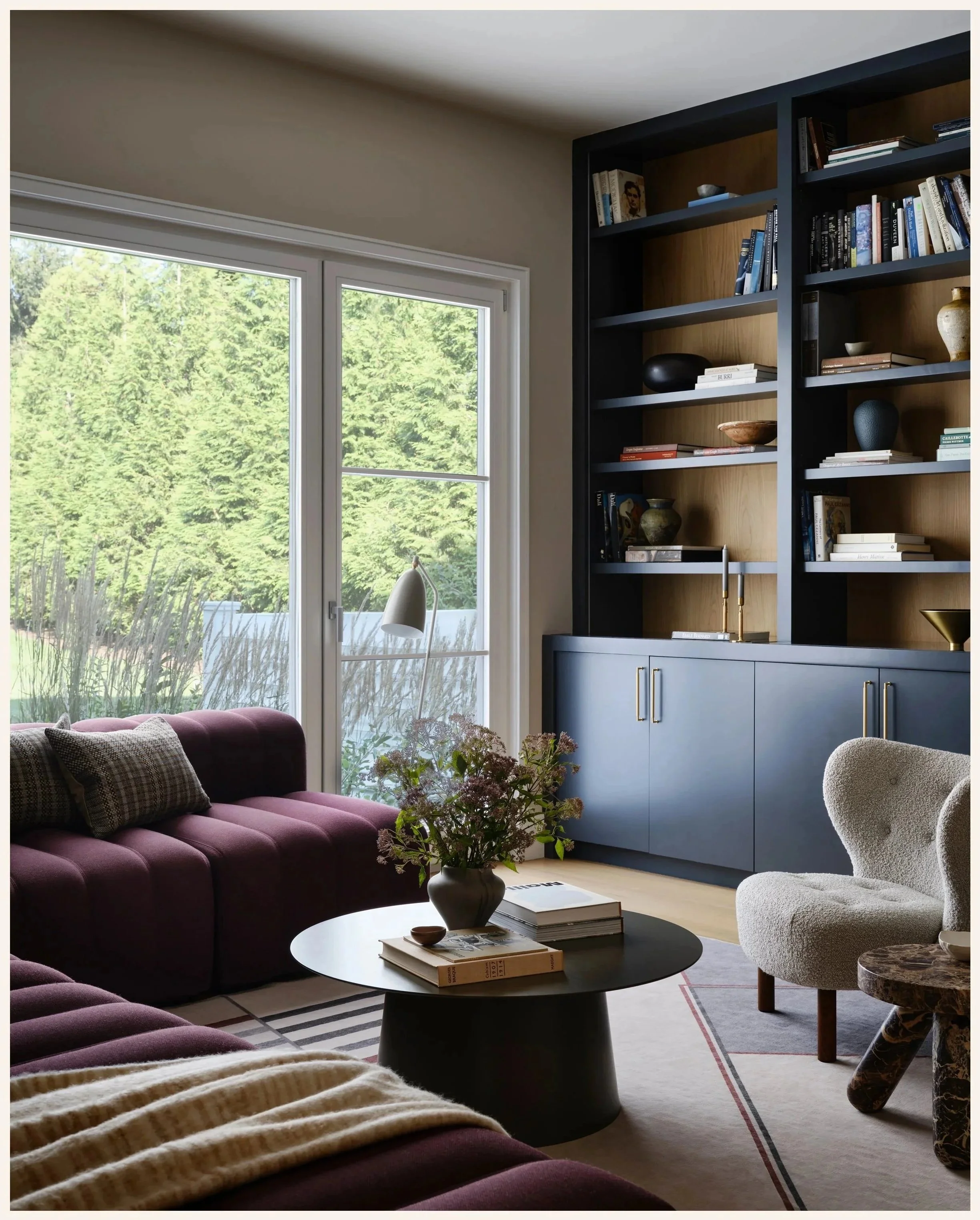 Living room with large window, purple sofa, white armchair, black coffee table with books and flowers, blue built-in bookshelf with decorative items, and a view of greenery outside.