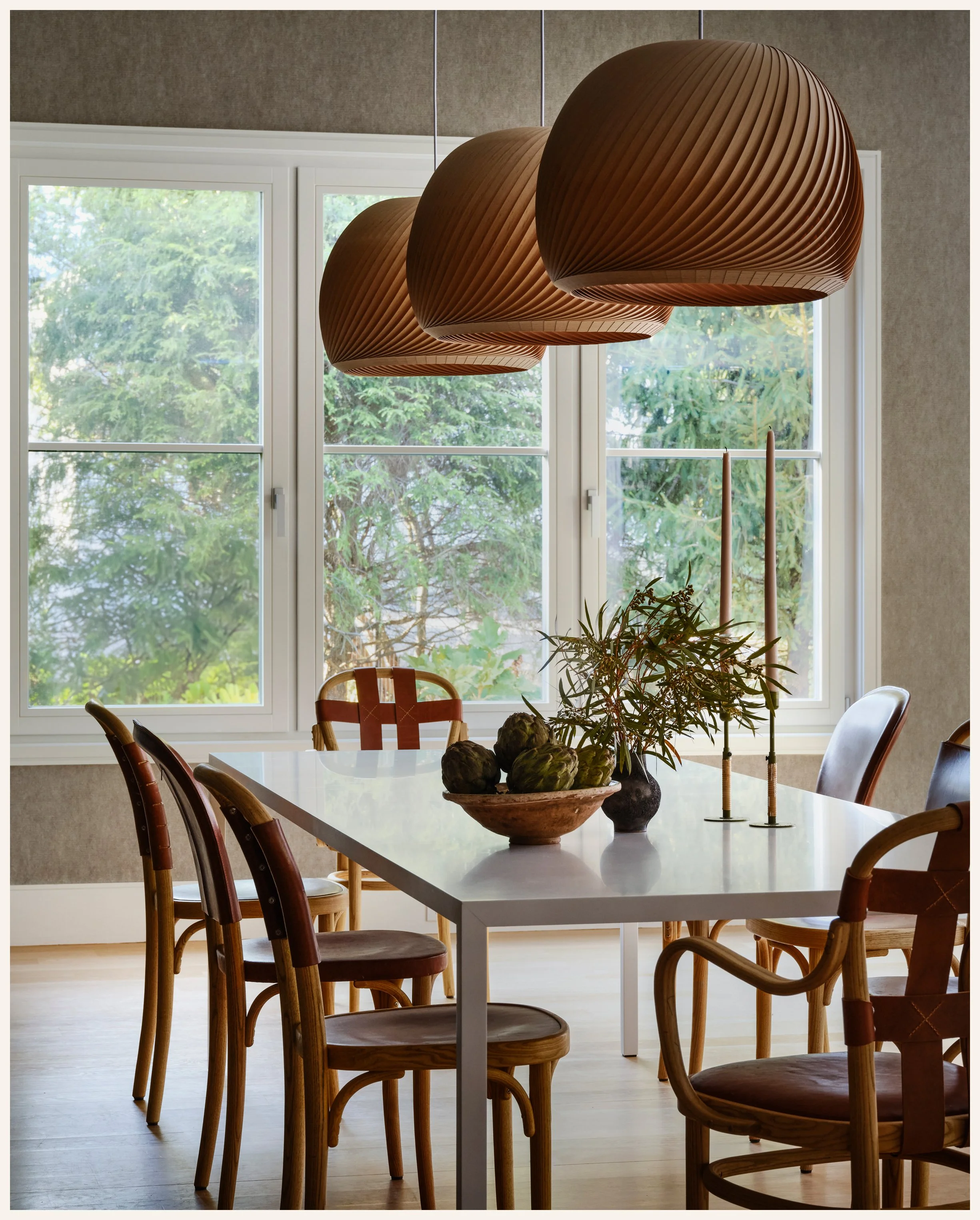 A dining room with a white table surrounded by wooden chairs, large window with a view of green trees outside, and three modern wooden pendant lights hanging above the table. The table has a decorative bowl with artichokes, a small vase with leafy br