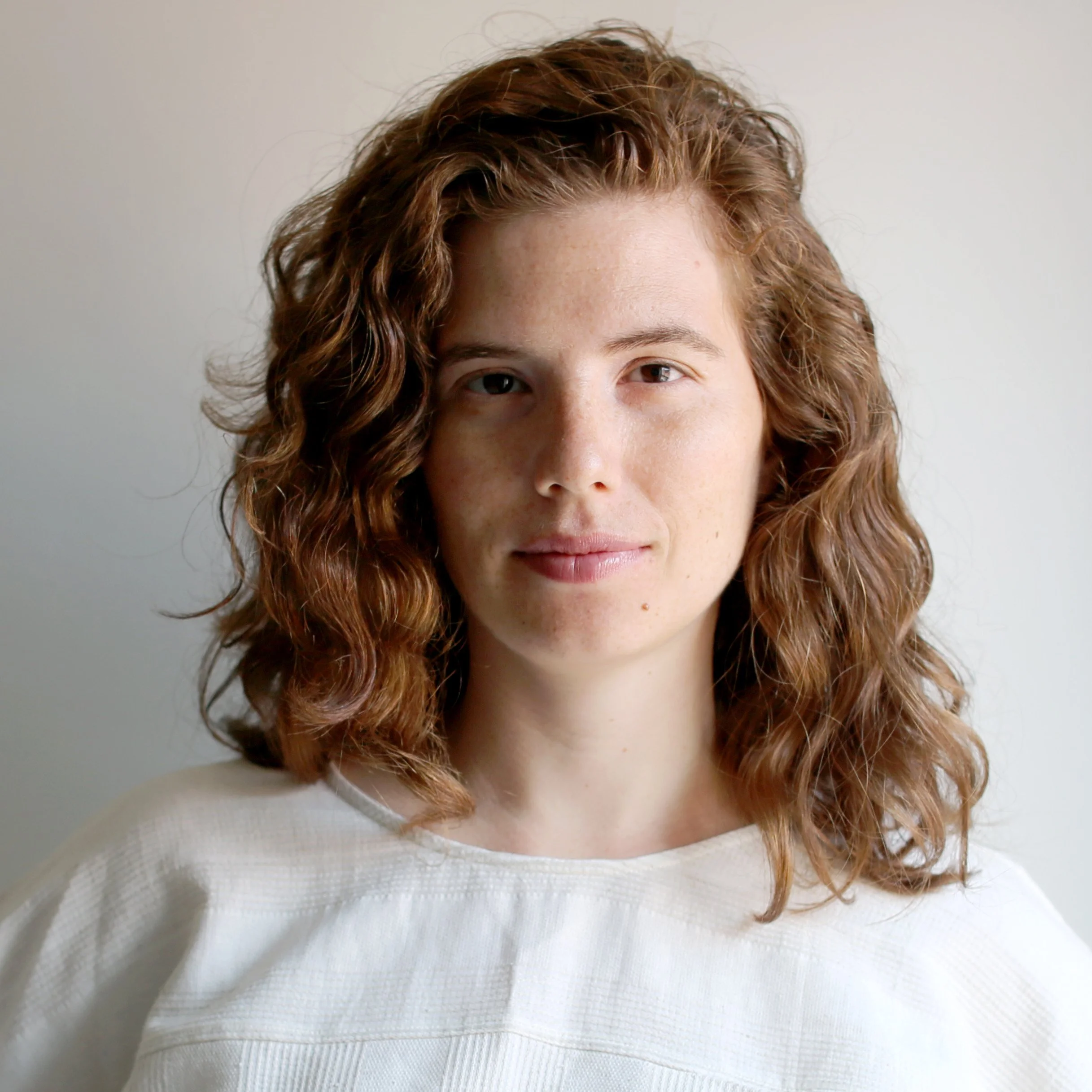 Portrait of a woman with curly, reddish-brown hair, wearing a white top, standing against a plain light-colored background.
