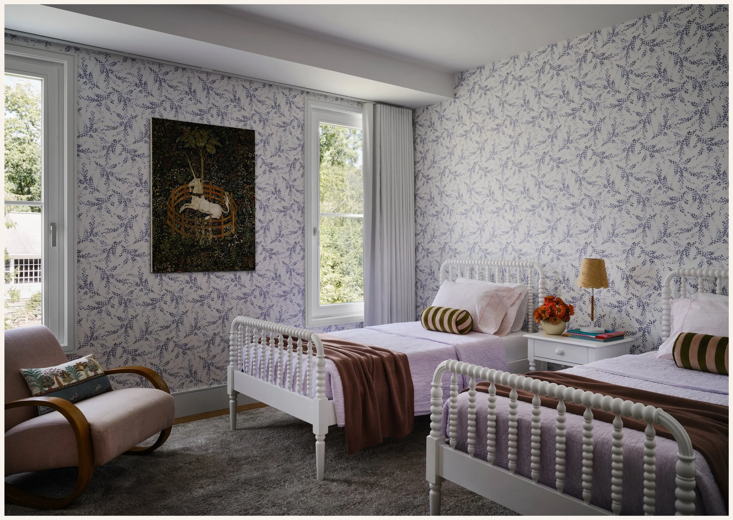 A bedroom with two twin beds with white metal frames, lavender bedding, and striped bolster pillows. A small white bedside table with a lamp, books, and a flower arrangement is between the beds. A pink armchair with a floral cushion is on the left. T