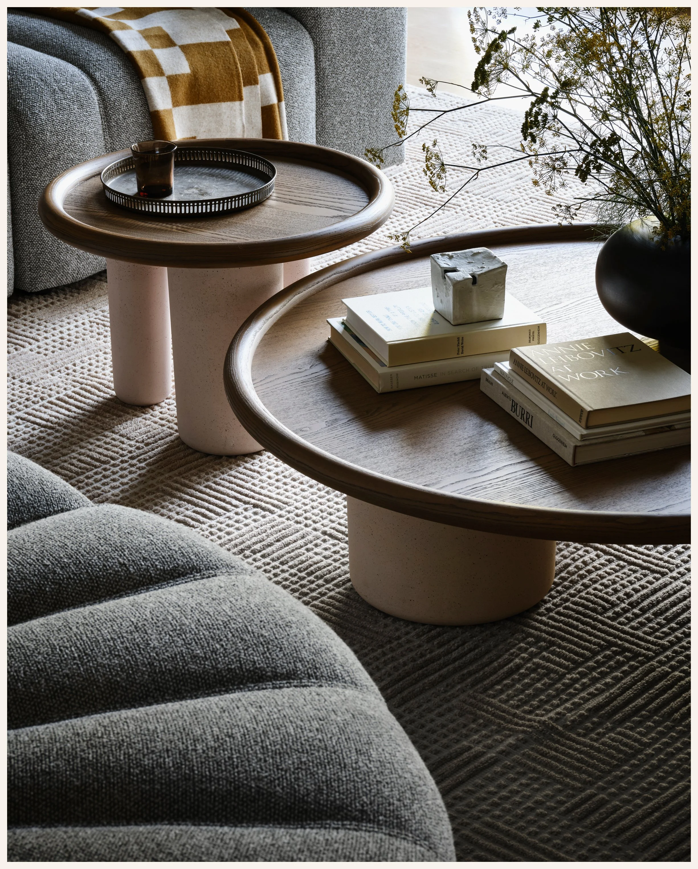 Living room with a textured gray sofa, round wooden coffee table, and a smaller matching side table. On the side table, there's a tray with a dark glass and a small cylindrical object. The coffee table has a stack of books, a small cube-shaped item, 