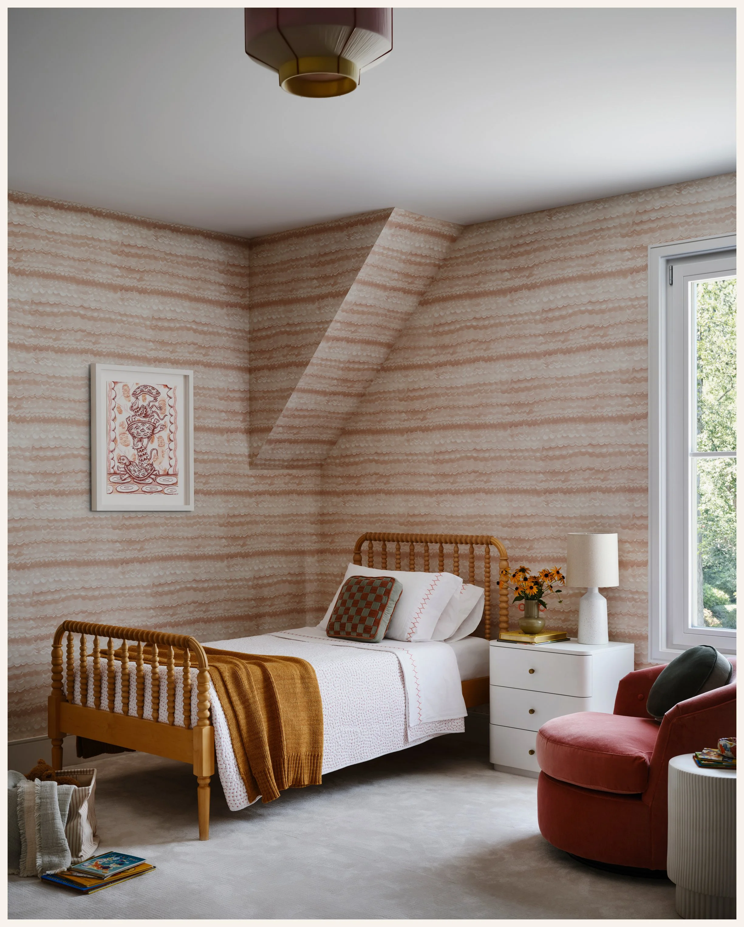 A bedroom with a wooden bed, white bedding, a decorative pillow, and a mustard-colored blanket. There is a white nightstand with a vase of flowers, a lamp, and some books. A pink velvet armchair with a dark throw pillow is next to the window. The roo