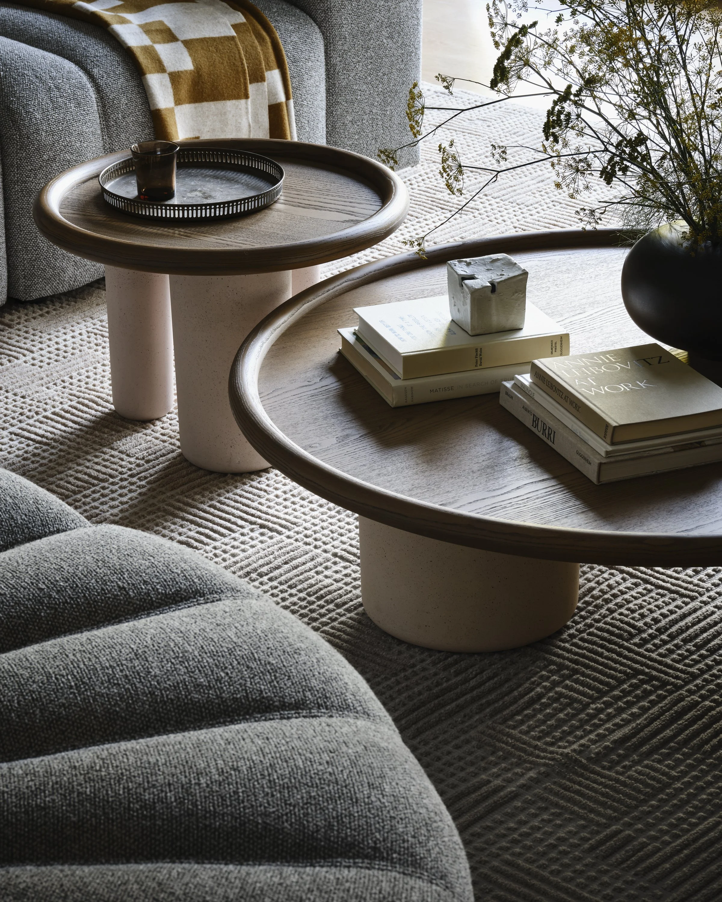 Living room detail with two round coffee tables, one with books and a ceramic object, another with a tray and a glass, a gray sofa with a yellow checkered blanket, a textured rug, a vase with flowers, a window with natural light, and a custom rug.