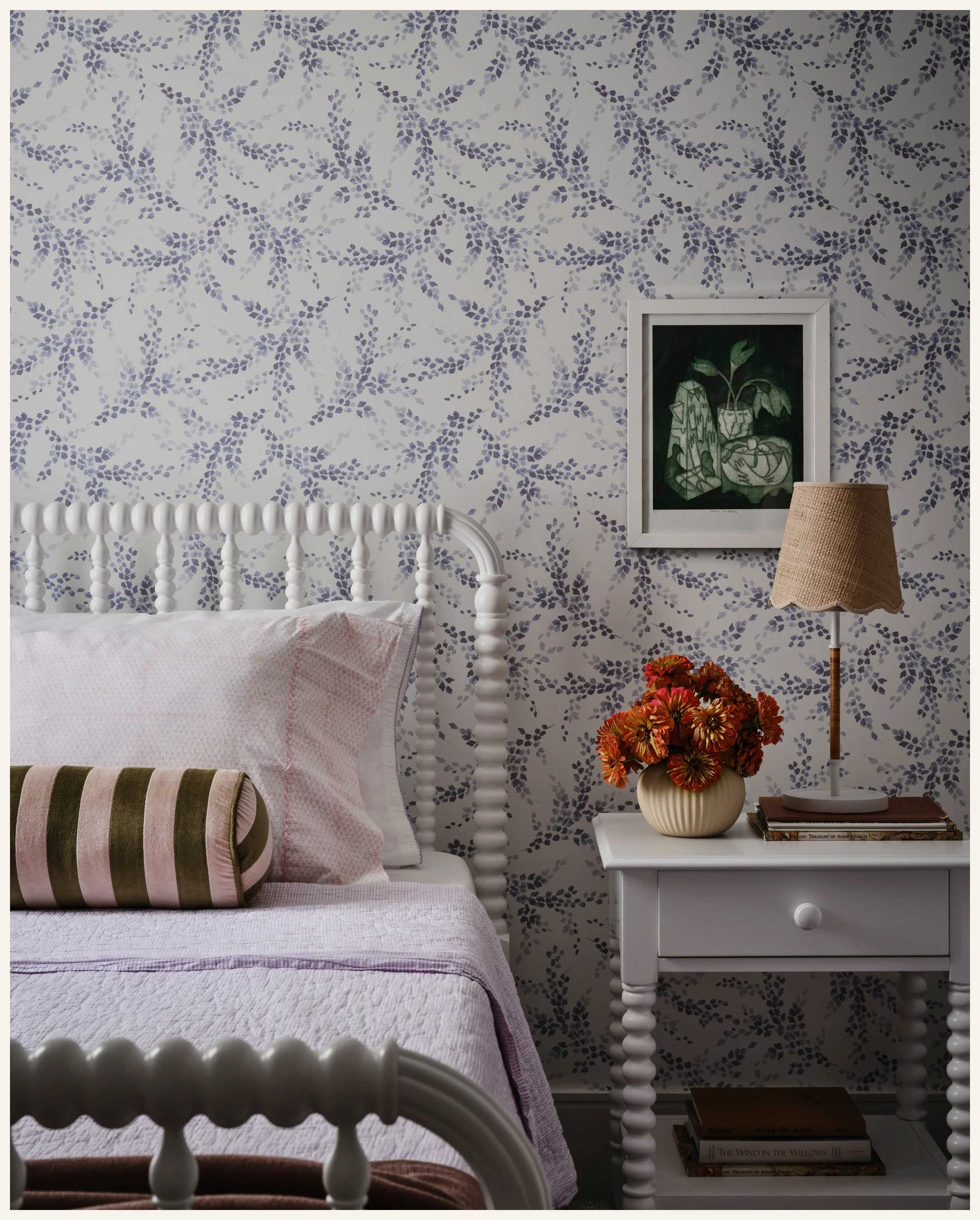 A bedroom scene featuring a white metal bed with purple and white bedding, a striped bolster pillow, a white bedside table with a beige lampshade, a vase of orange flowers, and framed artwork on the wall with a leaf pattern wallpaper.