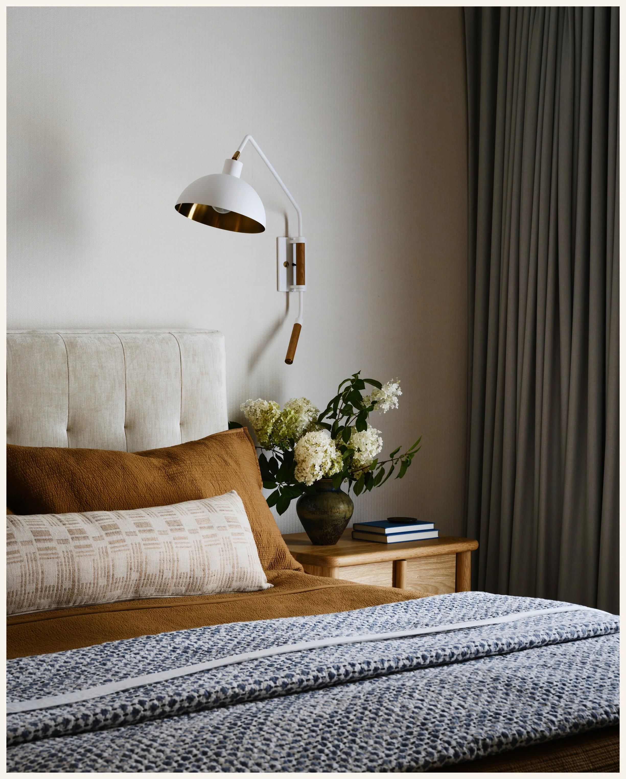 A bedroom with a beige upholstered headboard, a brown bedspread, and decorative pillows. There is a wooden nightstand with a vase of white flowers and a couple of books. A white wall-mounted lamp with gold accents is attached to the wall, and dark cu