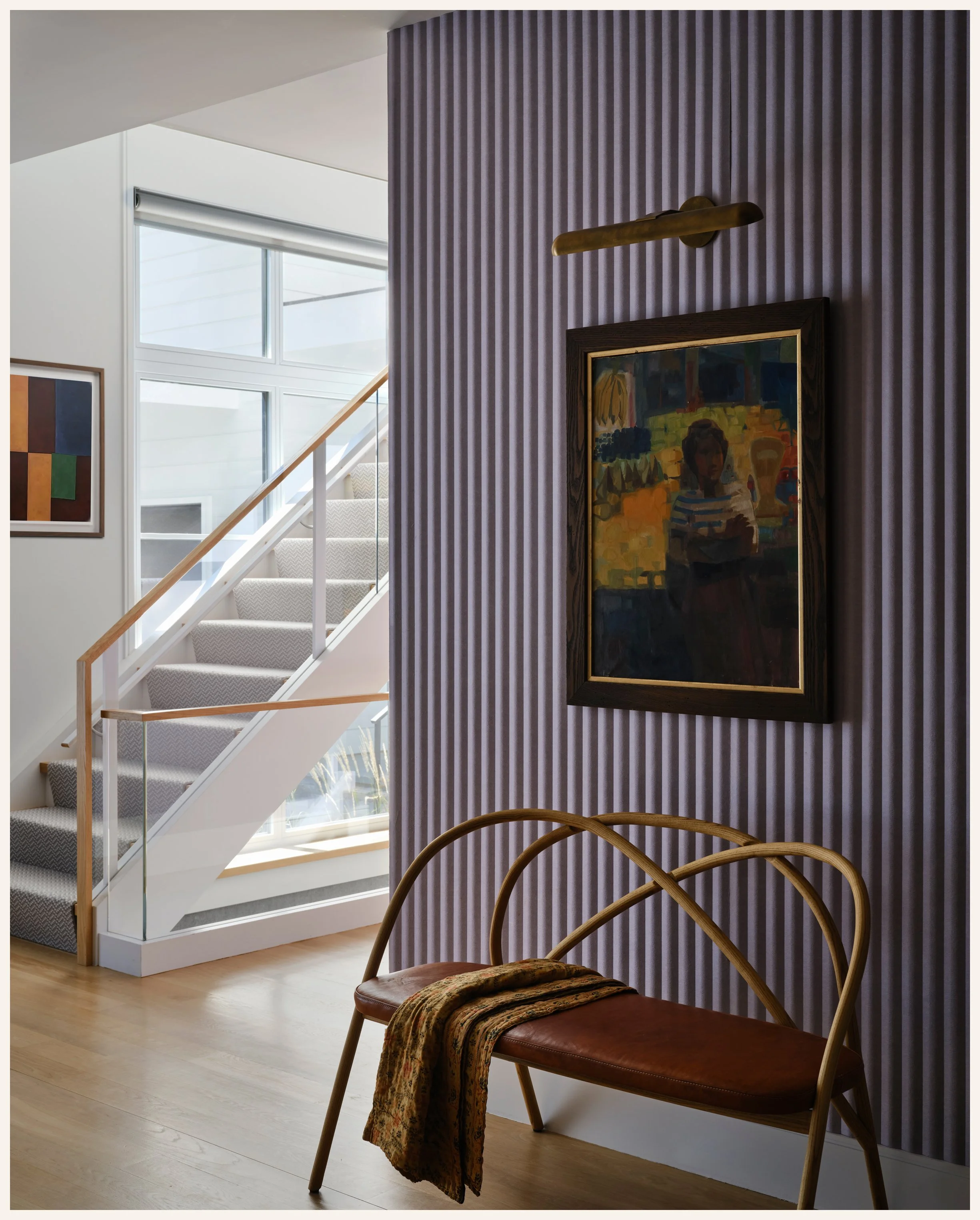 Interior of a modern home with a curved staircase, purple textured accent wall, framed artwork, wooden bench with a throw blanket, and large windows.