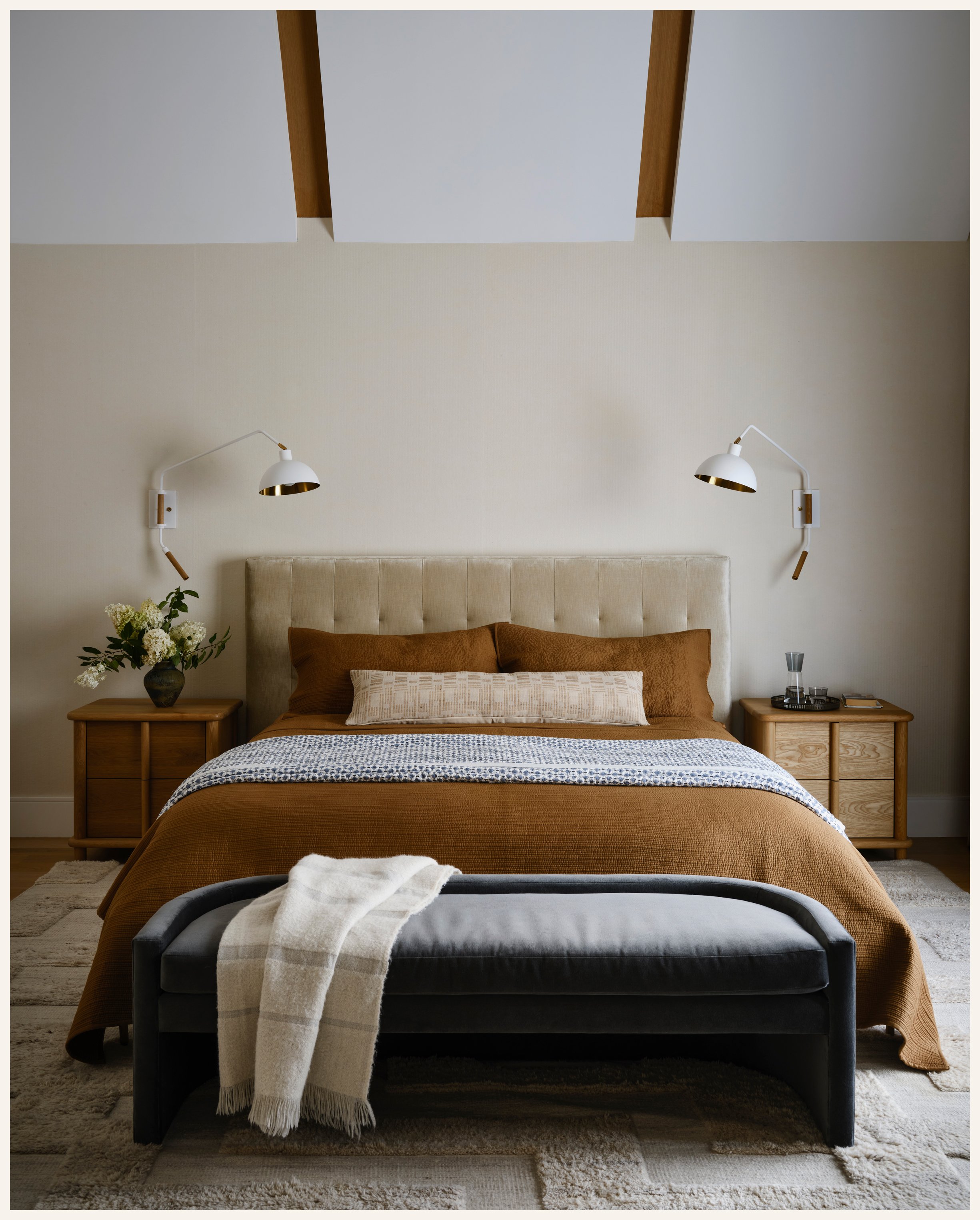 A bedroom with a beige headboard, brown bedding, and two wooden nightstands. Wall-mounted white lamps with gold accents are on each side. A bench with a blanket is at the foot of the bed, and a rug lies on the floor. There are flowers on the left nig