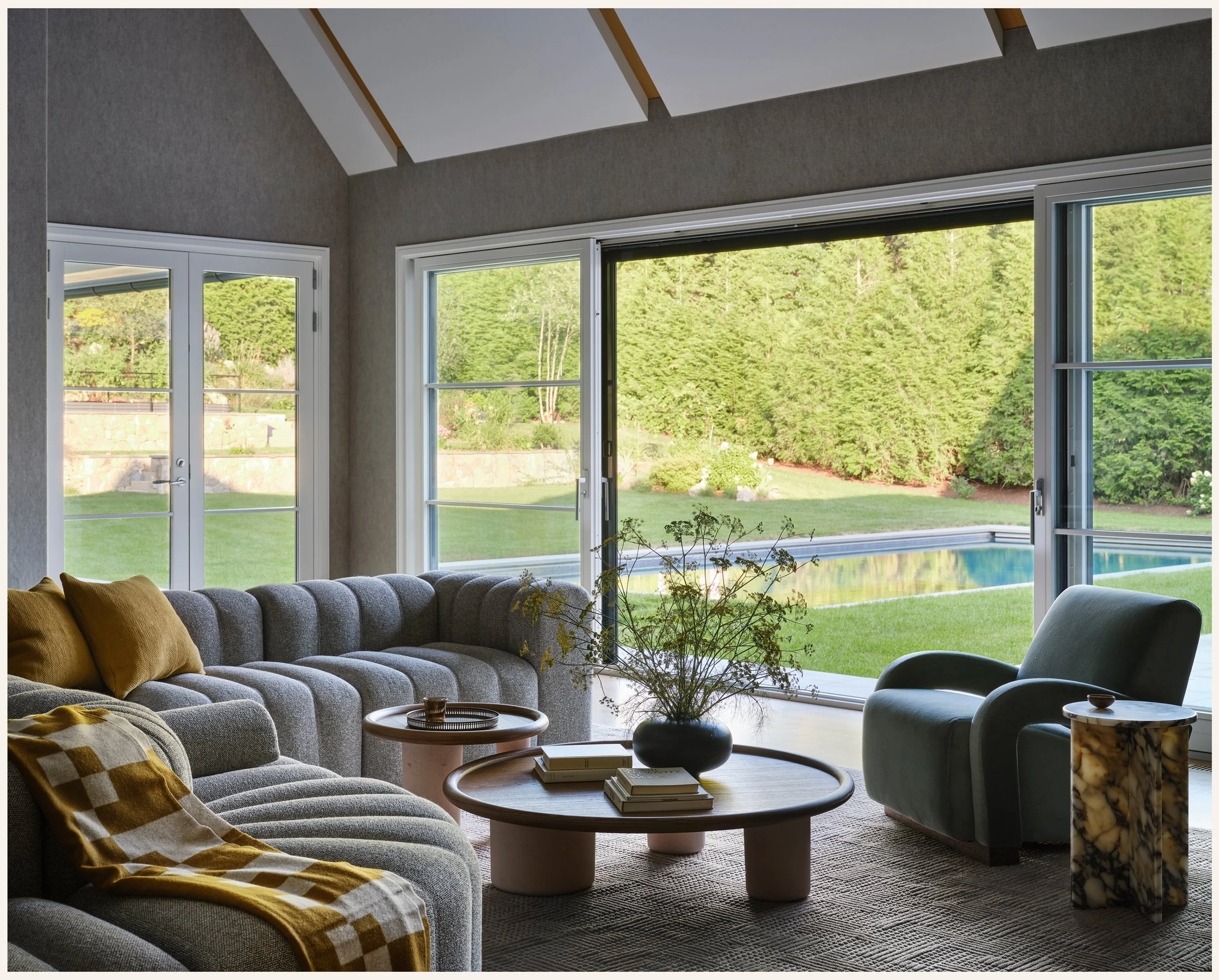 Living room with large sliding glass doors showing a backyard with a pool, green lawn, trees, and bushes, furnished with a gray sectional sofa, a light blue armchair, a round coffee table with a potted plant, books, and decorative pillows, and an abs