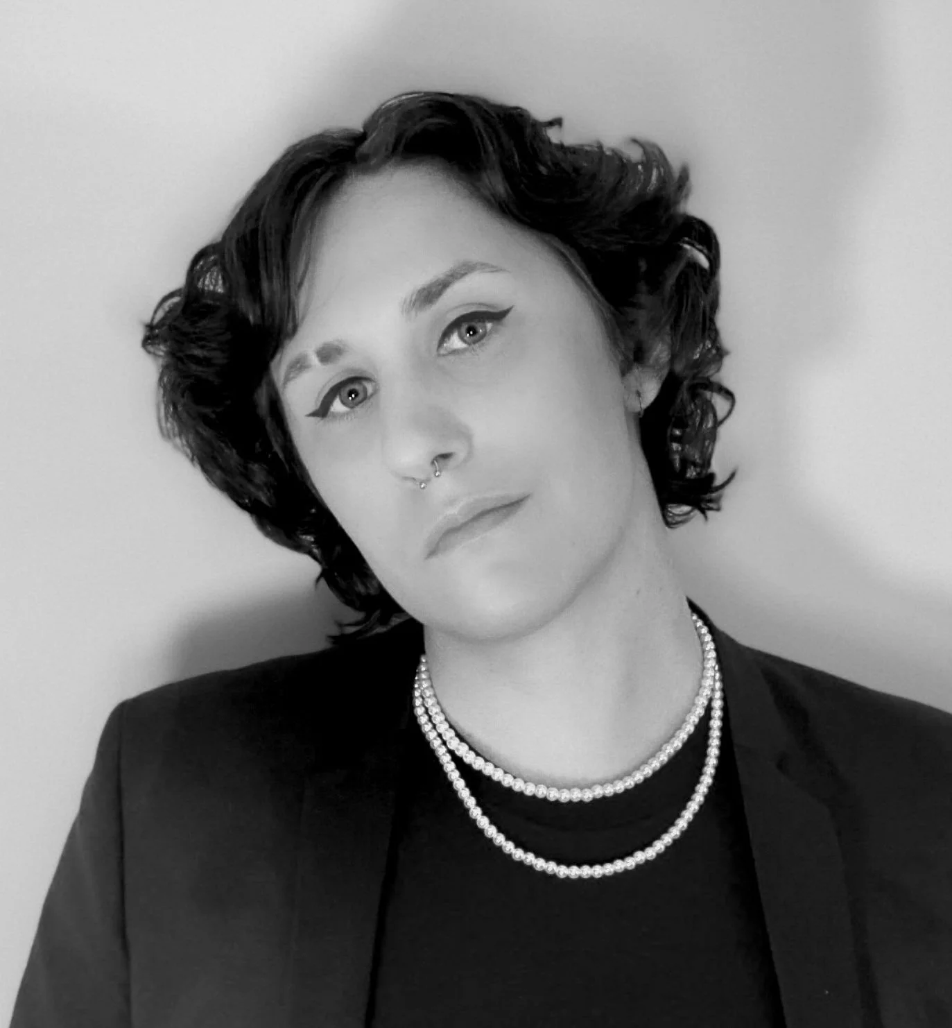 Close up black and white portrait of torrin, who is wearing a dark blazer and a double strand of pearls. She is looking directly into the camera and her head is tilted slightly to the right. They have short wavy hair.