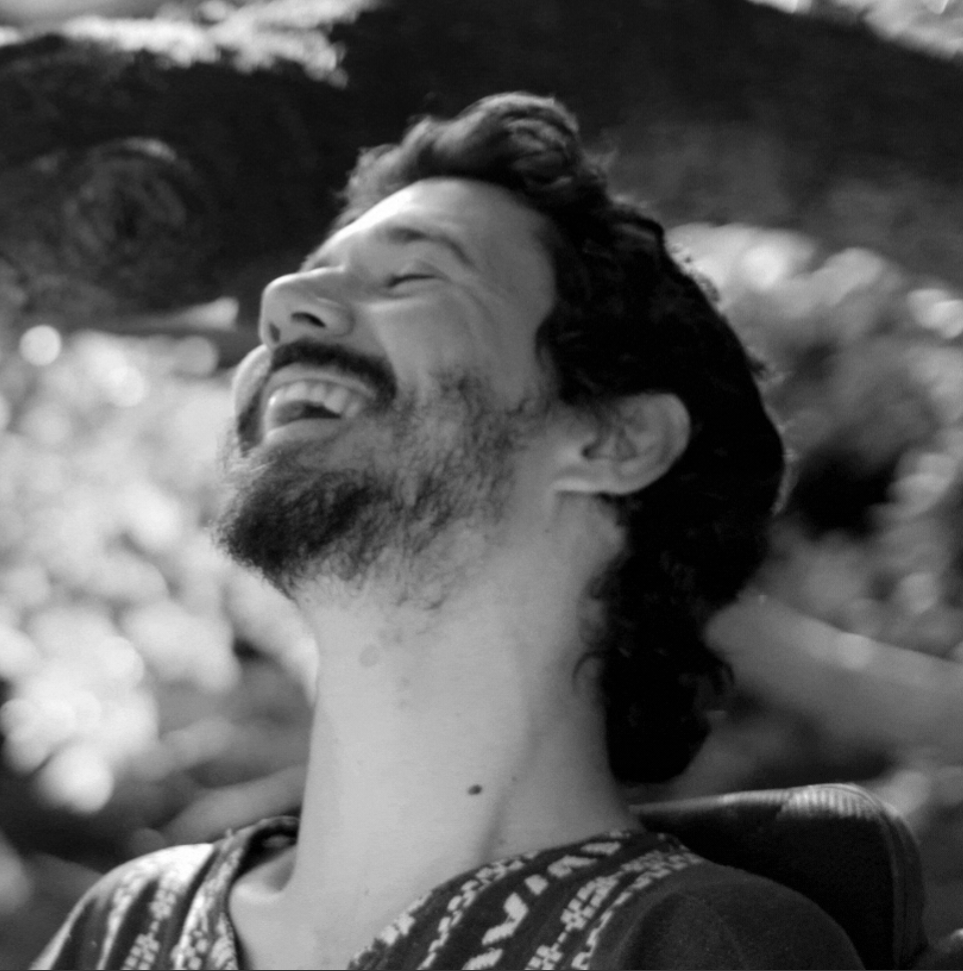 Black and white close up photo of Ekiwah. His head is tilted backward and he has a large smile on his face. Ekiwah has a beard and dark curly hair.