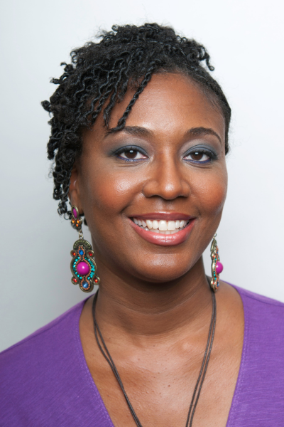 Close up photo of Camisha, a Black cis-woman with gingerbread brown skin. Flat twists in her hair frame her face. She is wearing a v-neck purple dress, bright jewel tone earrings, and dark teal eye shadow.