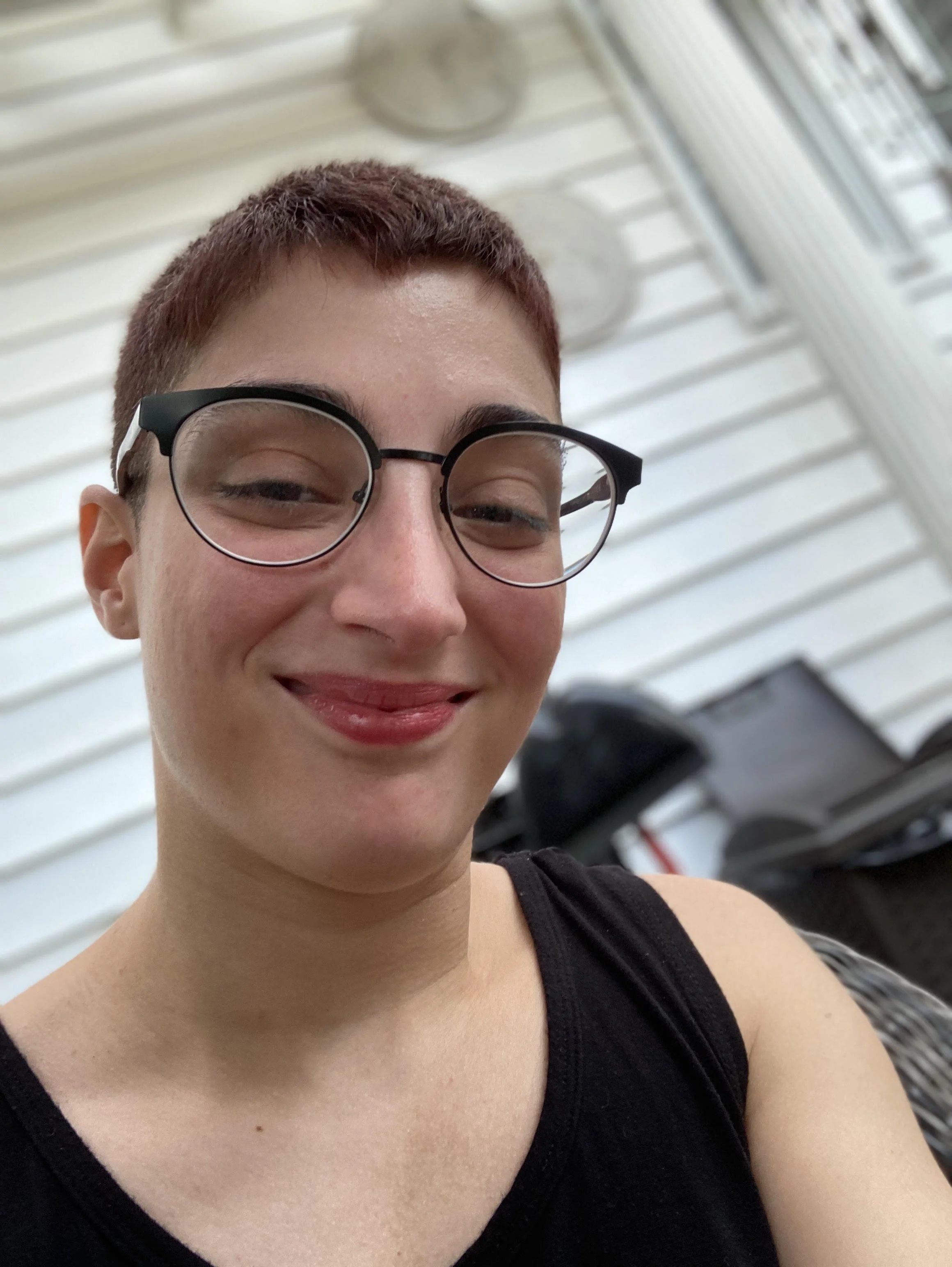 Photo is a close up of Liv seated inside with a computer in the background. She has very short brown hair and glasses and is wearing a black tank top. Liv is smiling and looking directly at the camera.
