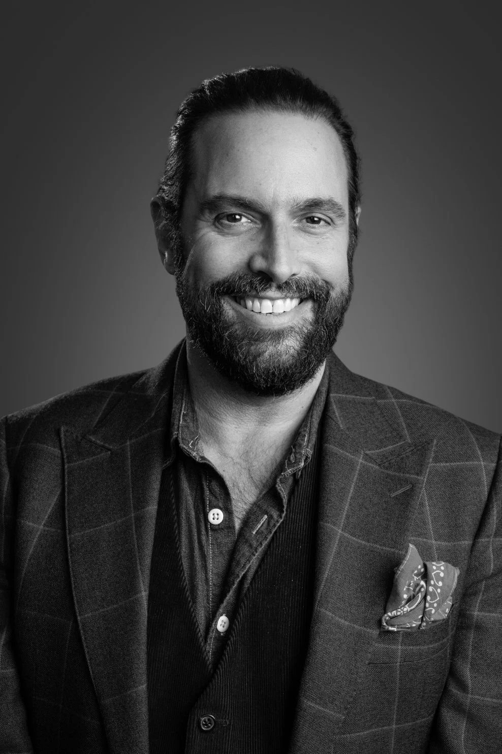 A smiling man with dark hair and a beard, wearing a checkered blazer and a pocket square, in a black and white professional portrait.