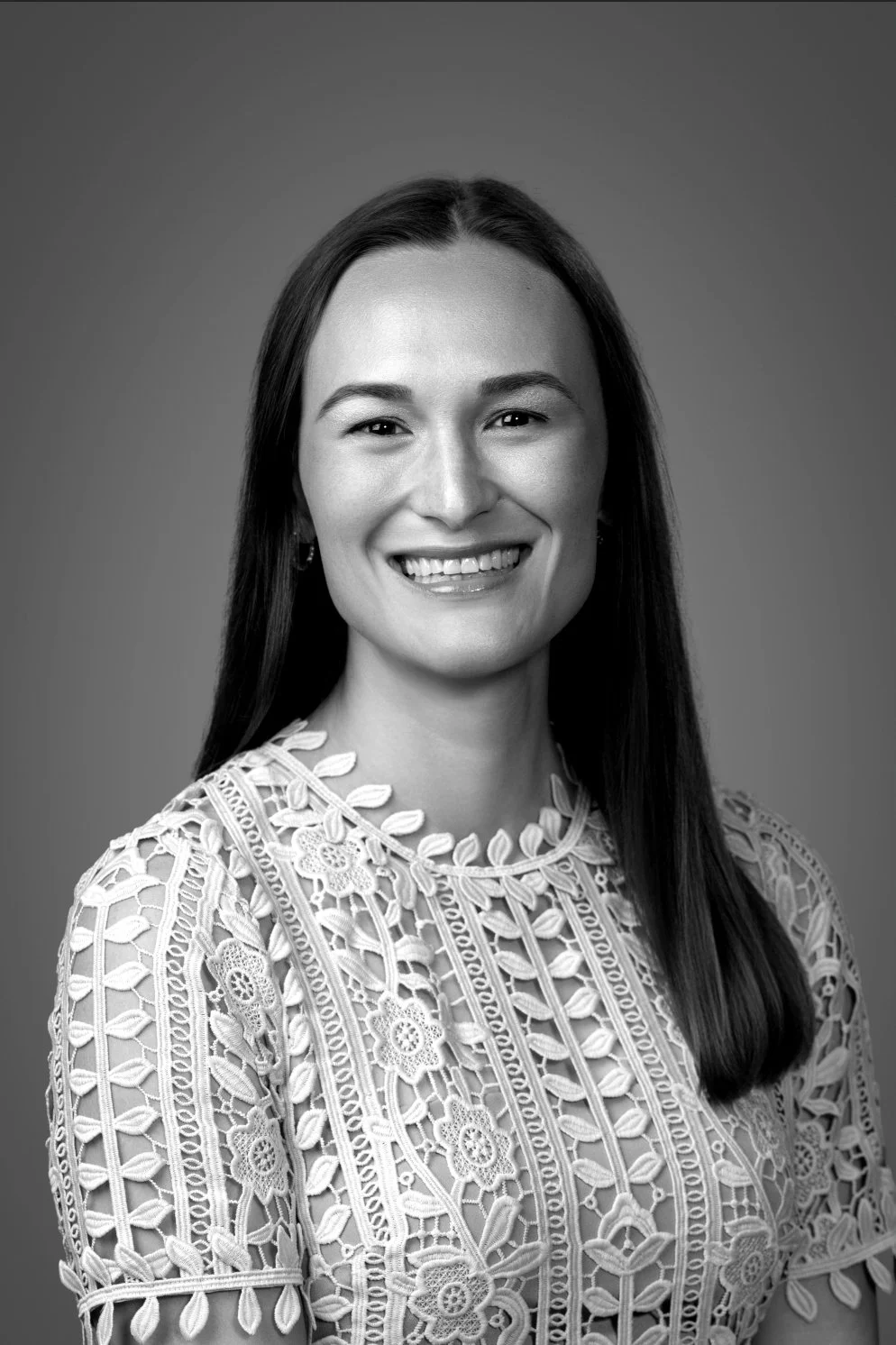 Black and white portrait of a smiling woman with long dark hair, wearing a detailed lace top, against a plain background.