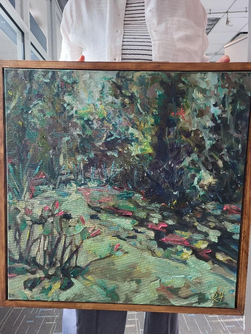 A person holding a framed landscape painting of a pond with lily pads and surrounding trees, in an indoor setting.