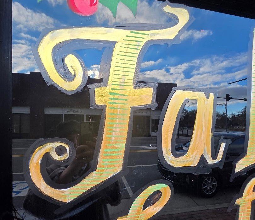 Close-up of a painted sign with large, decorative yellow and orange letters reflecting in a window, with a street scene and cloudy sky in the background.
