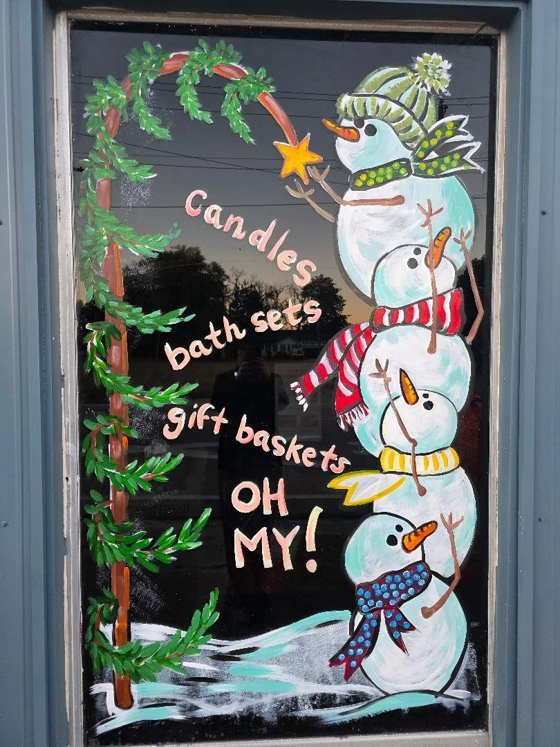 Decorative window with a painted snowman and snowballs wearing a scarf, hat, and earmuffs, surrounded by green evergreen branches. The window features pink lettering that reads, "Candles, bath sets, gift baskets, OH MY!"