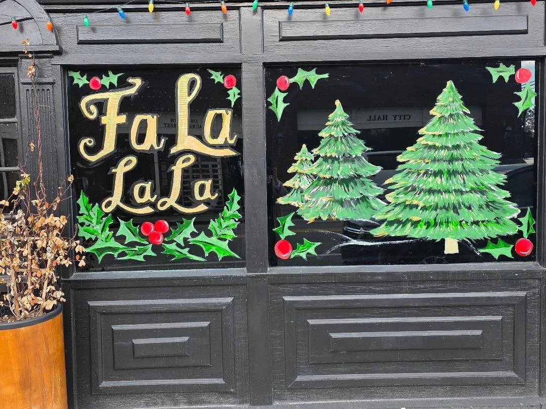Decorated storefront window with Christmas-themed decorations, including pine trees and holly leaves with red berries, and the phrase "Fa La La" in festive gold lettering.