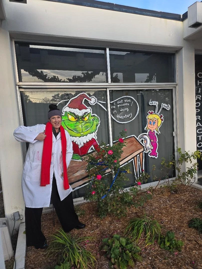 Grinch and a girl with candy cane antennae. The window has a painted Santa hat on the Grinch and a speech bubble saying, 'don't steal the presents! I'm doing inventory.' There are pink roses and other plants outside the window.