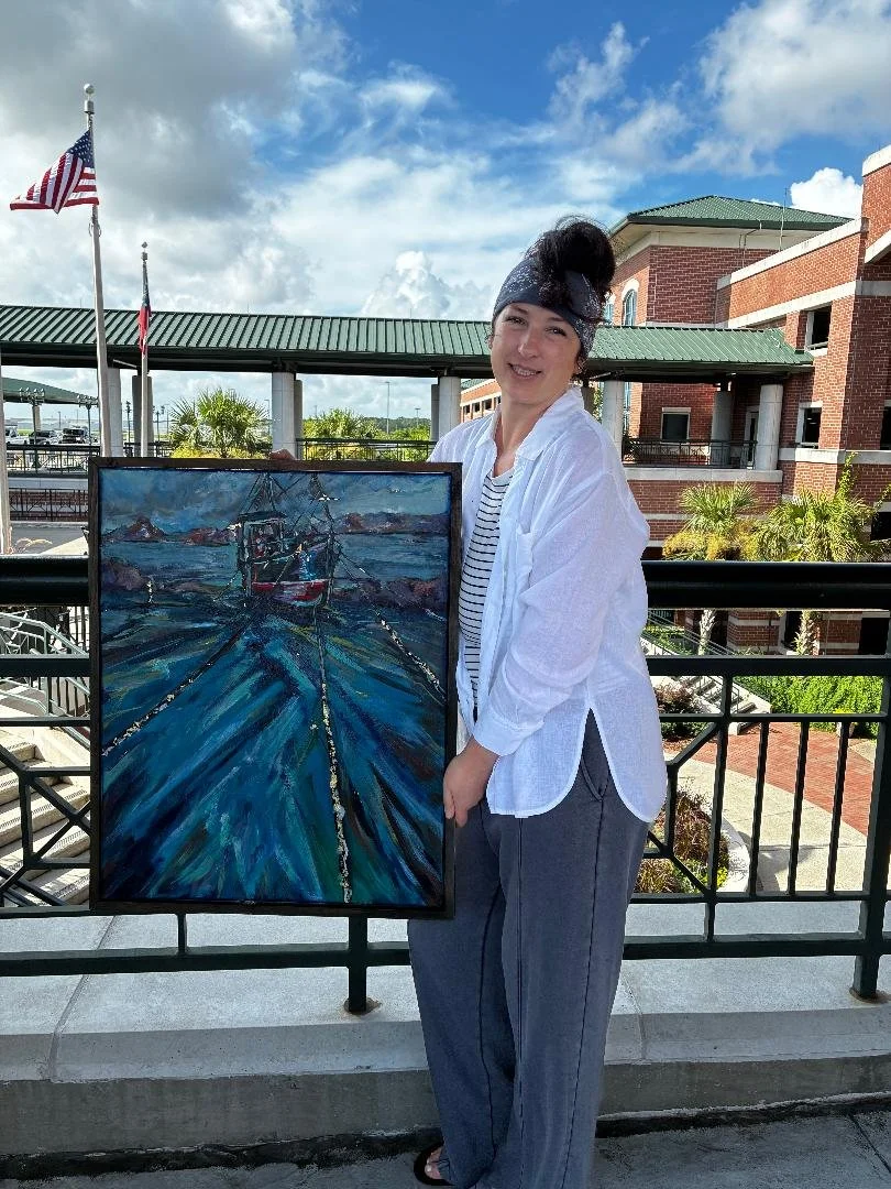 A woman standing outdoors on a balcony, holding a painting of a dock with boats on water under a cloudy sky, with a building and trees in the background.
