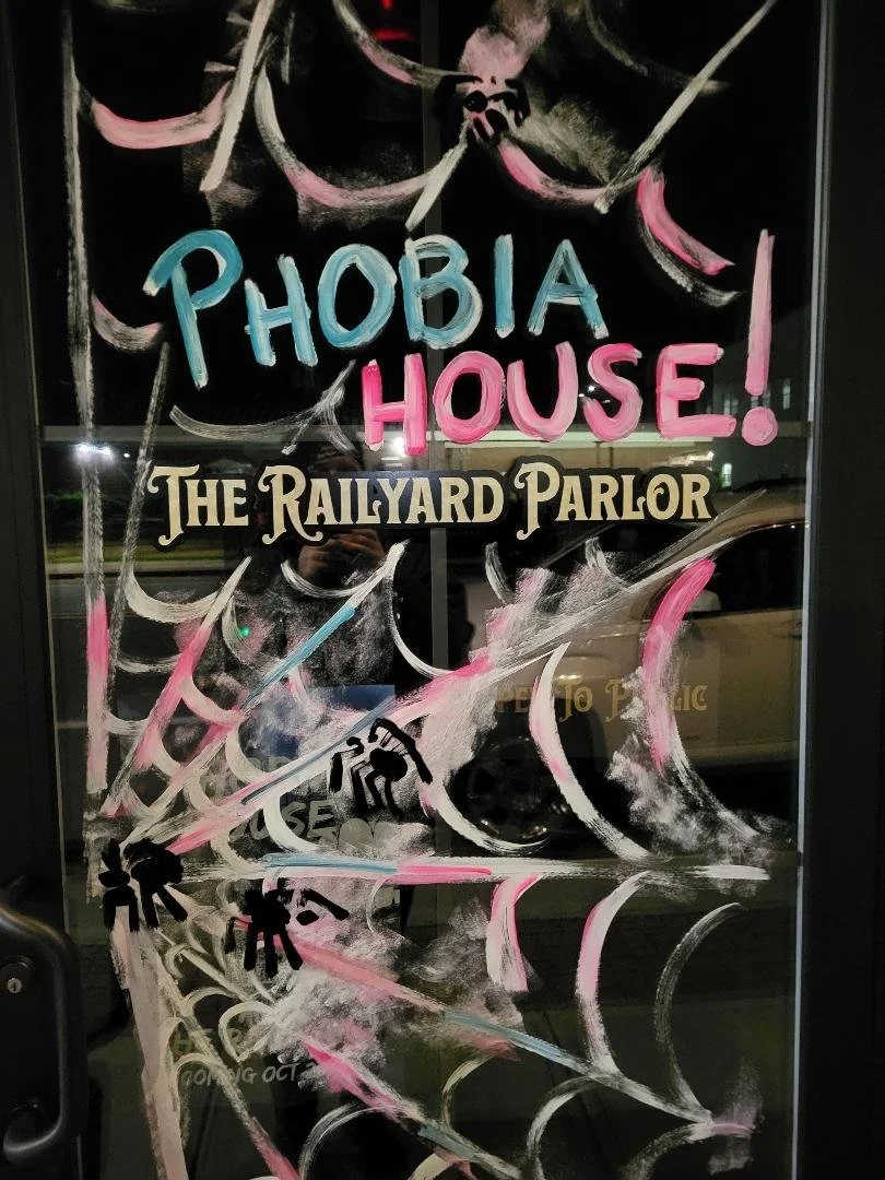 A storefront window with hand-painted pink and white spiderwebs and black spider illustrations, promoting 'Phobia House! The Railyard Parlor' with colorful text.